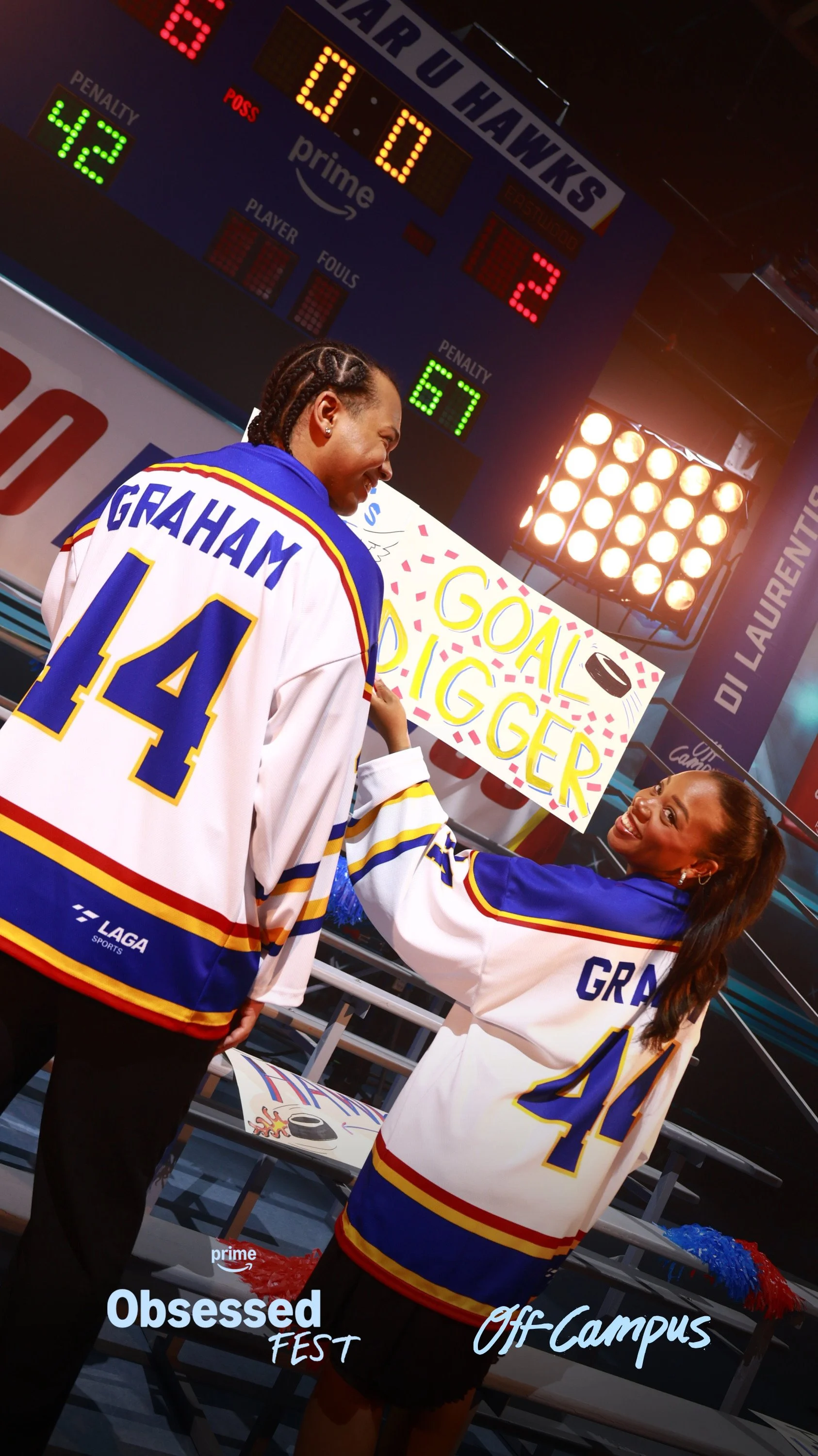 Two women wearing hockey jerseys are at an ice rink, holding a sign that says 'Goal Digger' with a drawn hockey puck. They are smiling and appear to be celebrating, with a hockey scoreboard in the background showing a tied score of 0-0. The scene is 