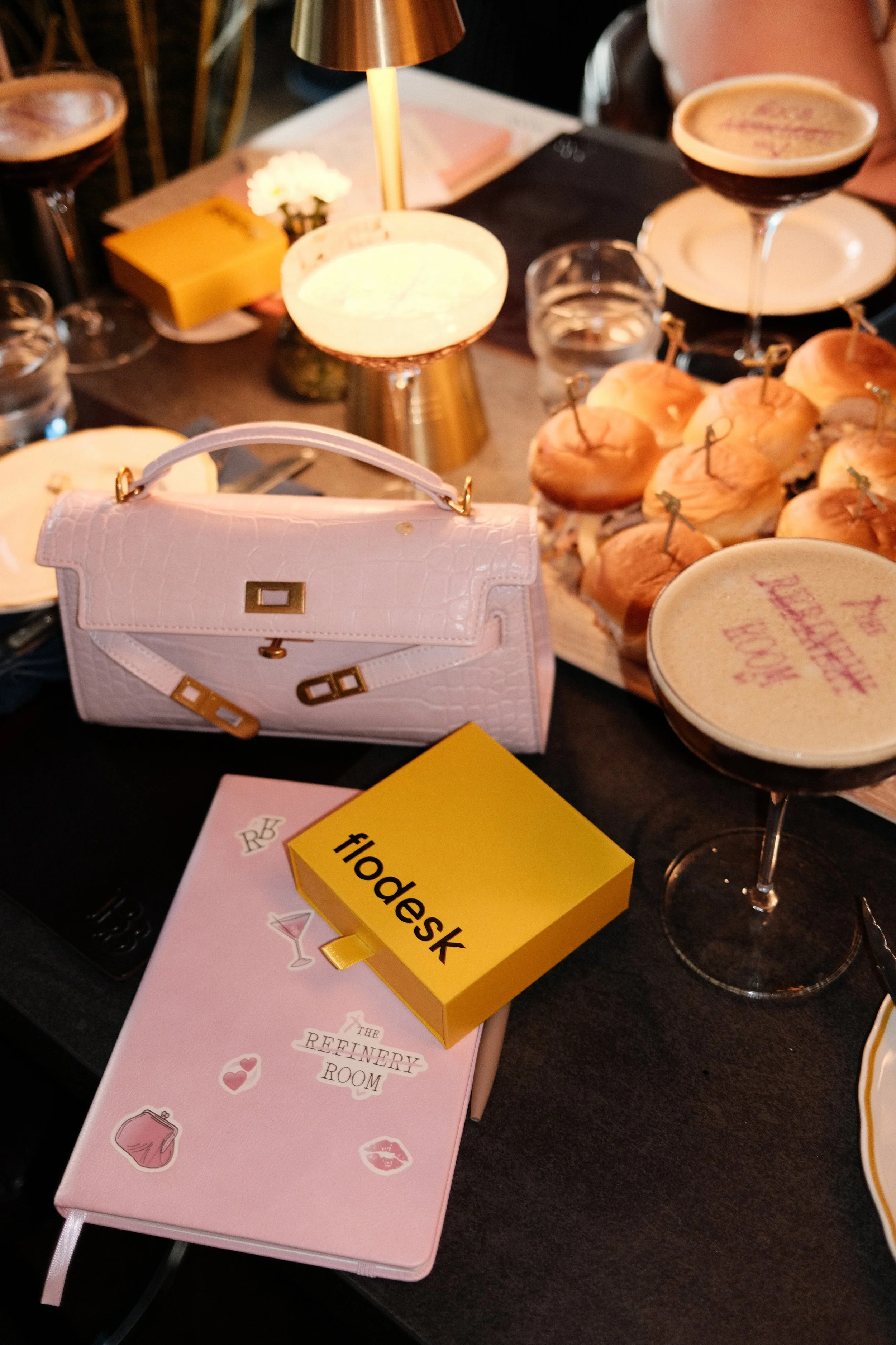A table with a pink notebook titled 'The Refinery Room', a yellow box labeled 'floedesk', two pink purses, a small vase with white flowers, a black menu, glasses of water, and several cocktails on stands.