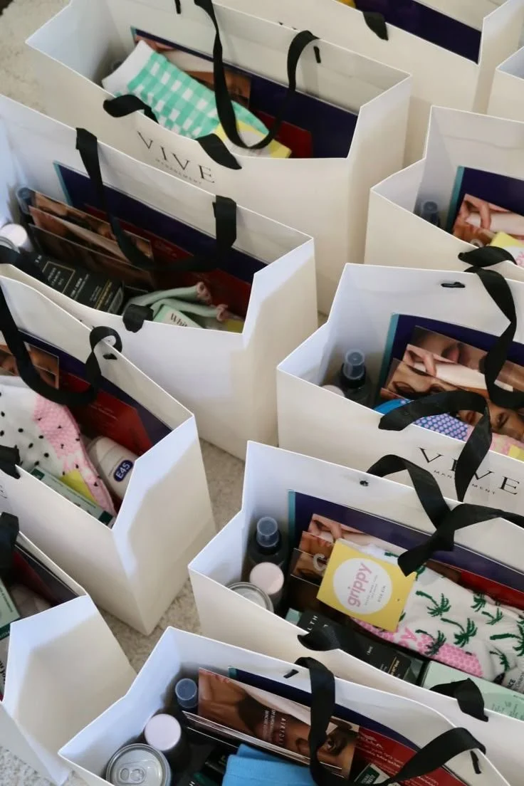 Multiple white shopping bags with black handles filled with various personal care products and gifts, placed on a carpeted floor.