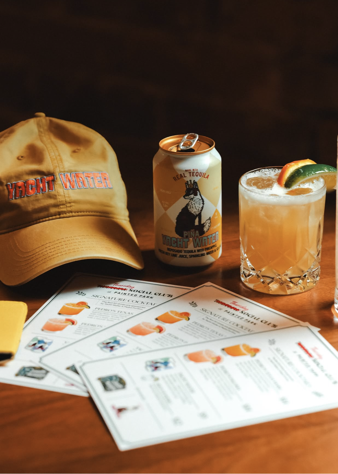 Yellow cap with red and blue embroidery reading 'Yacht Water', can of Piña Yacht Water cocktail, glass of cocktail garnished with lime and orange slices, social club menu on table.