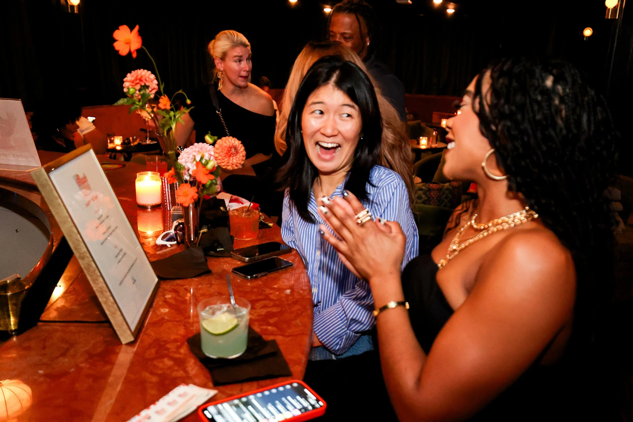 A group of women socializing at a bar or restaurant, smiling and engaging in conversation, with drinks and flowers on the table.
