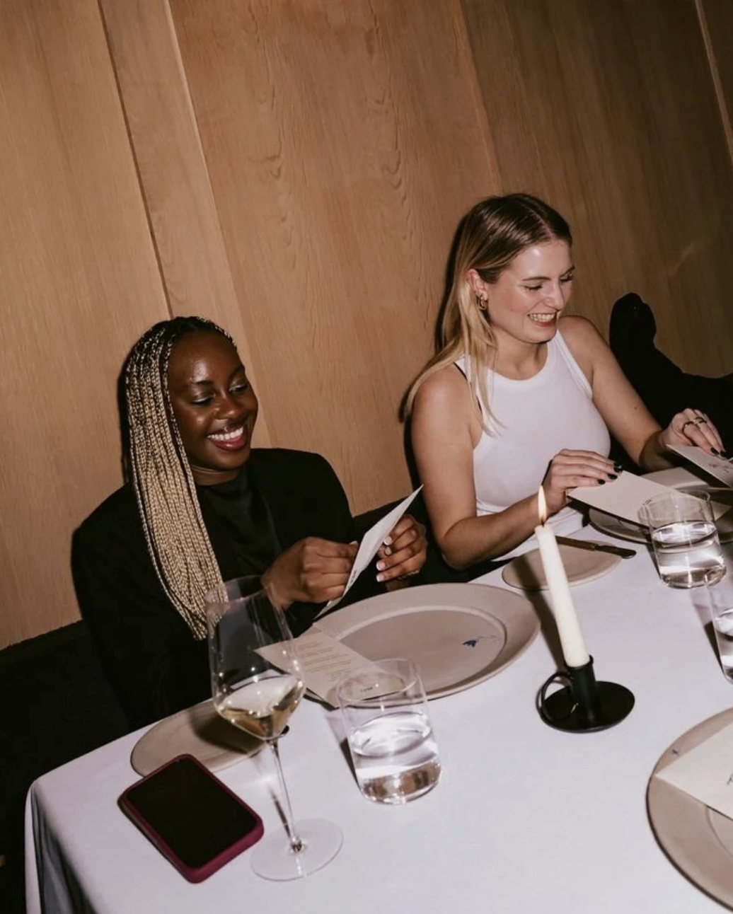 Two women sitting at a dining table, smiling and reading menus, with glasses of water, a glass of wine, a candle, and a phone in front of them.