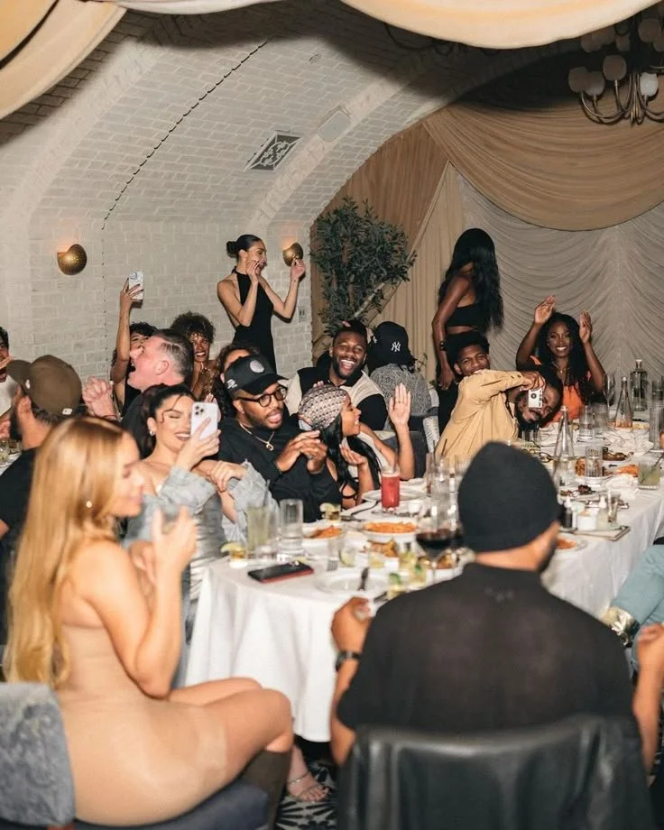 A diverse group of people enjoying a celebration at a banquet table with food and drinks, some are taking pictures, smiling, and clapping in a decorated indoor venue.