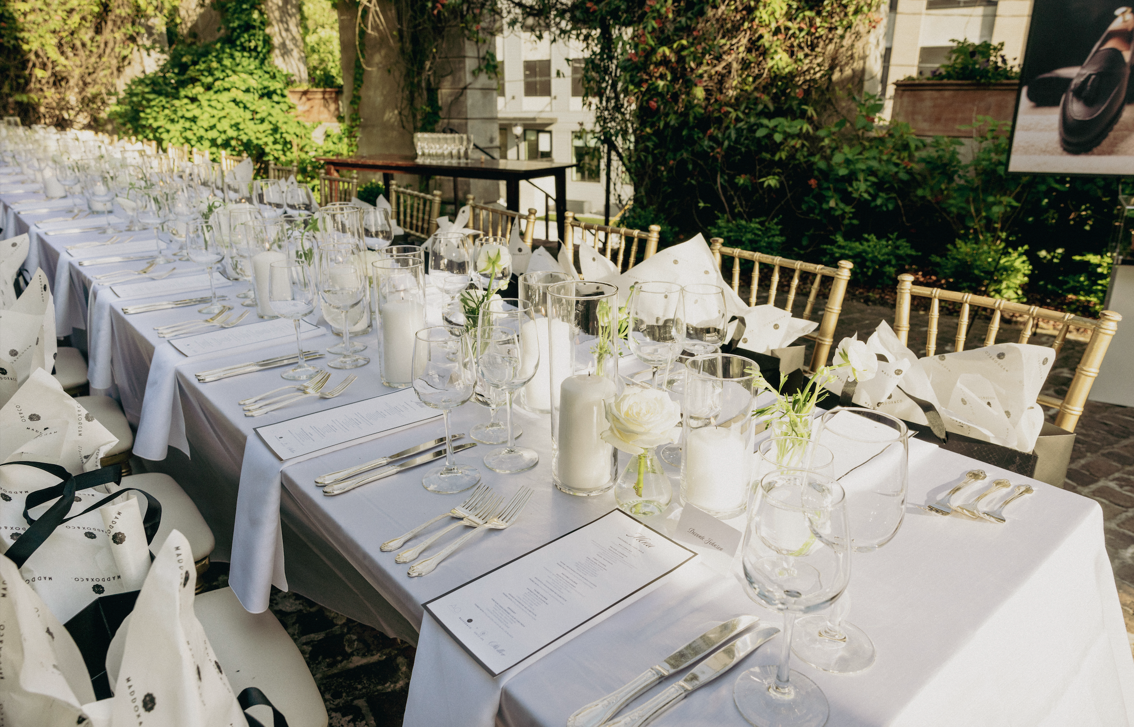 Elegant outdoor dining table set with white tablecloths, glassware, white napkins, and floral centerpieces on a patio with greenery.