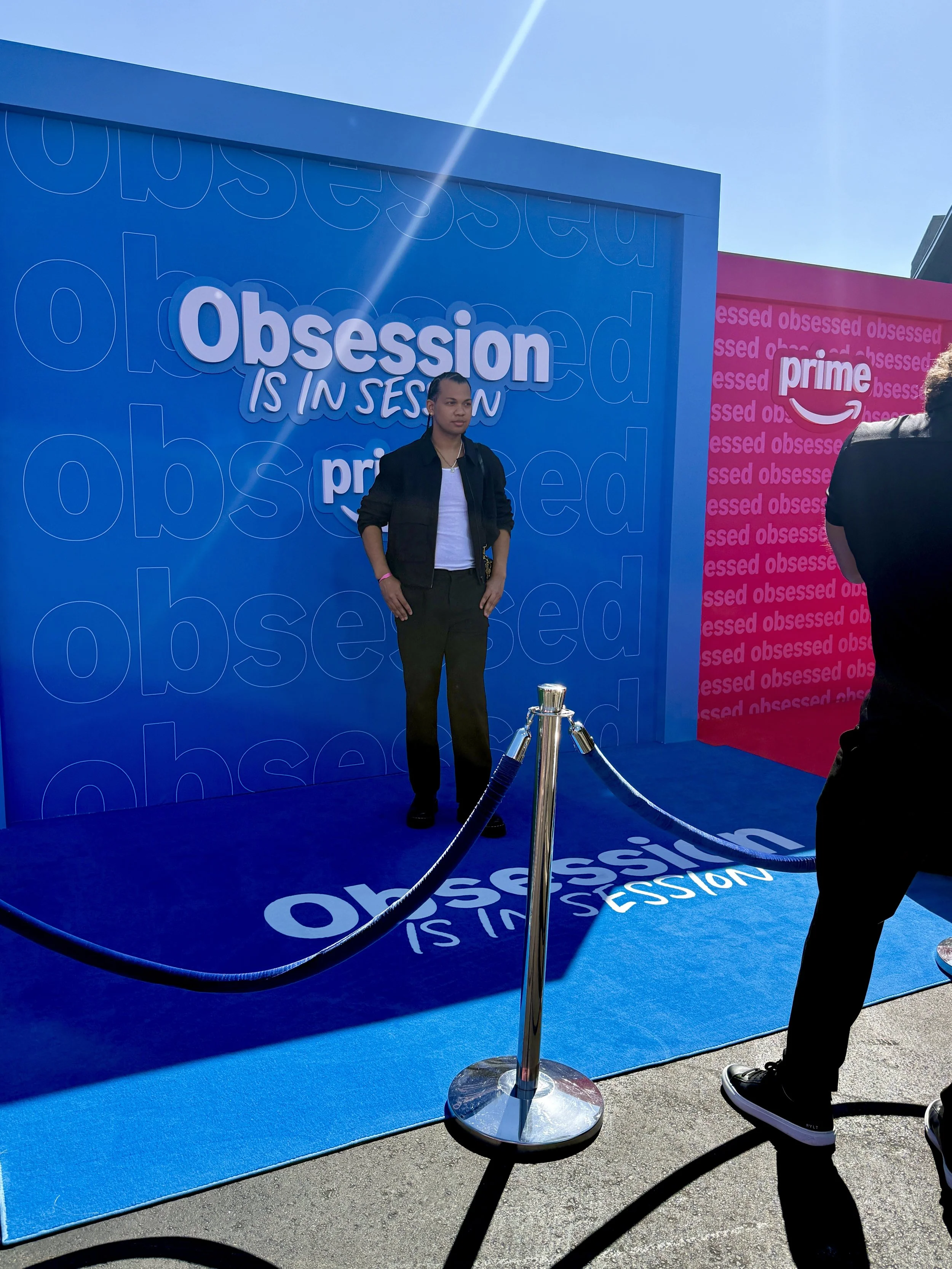 Person standing on a blue carpet with the words 'Obsession is in session' and 'Prime' logo on a backdrop, with a velvet rope barrier in front.