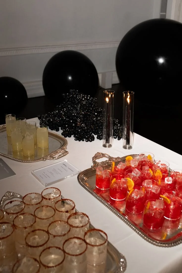 Table with three types of drinks in glasses, black balloons as decoration, candles, and black beads in the background.