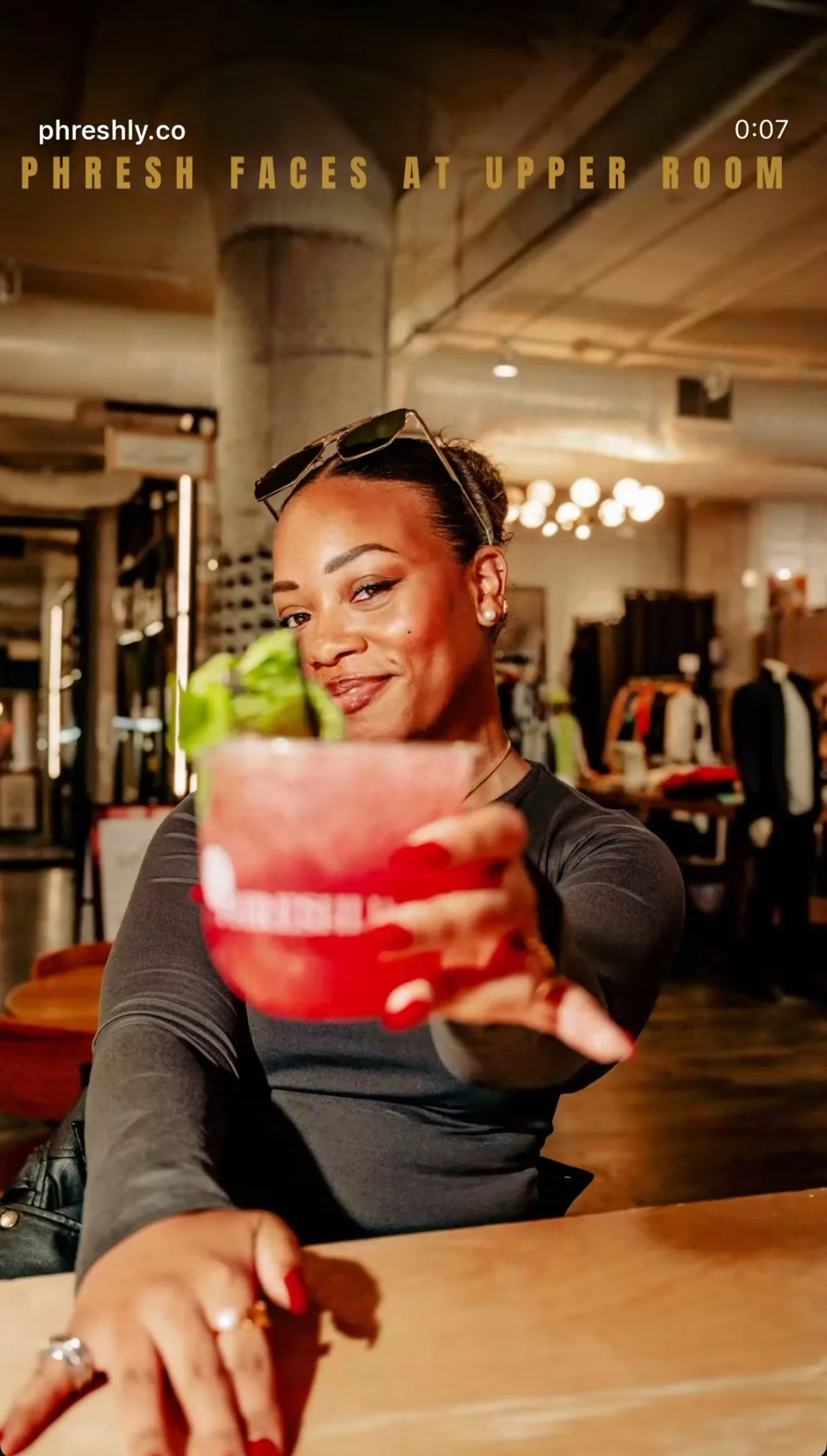A woman smiling and holding out a red cocktail garnished with a slice of cucumber, sitting in a well-lit indoor setting that appears to be a bar or restaurant.