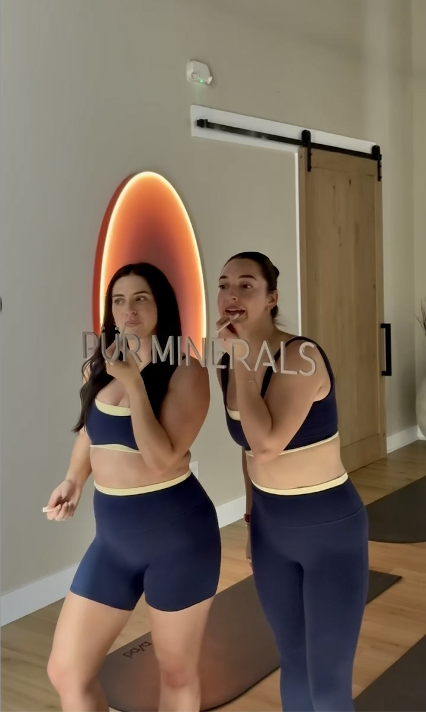 Two women in workout clothes standing in front of a mirror with the word 'PURMINERALS'. One woman is applying lipstick, and the other is applying makeup or lip gloss.