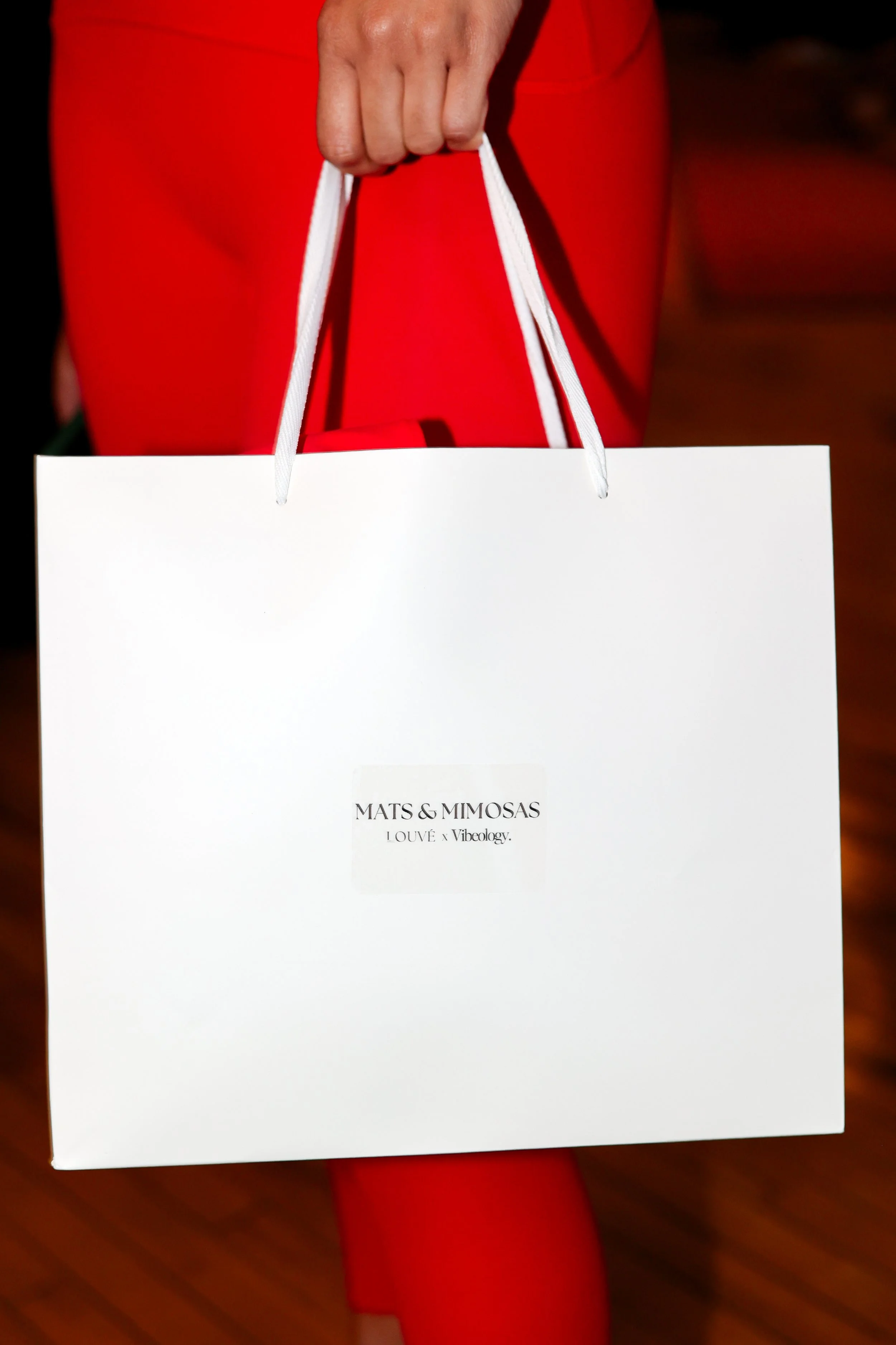 Person holding a white shopping bag with a label that reads 'MATS & MIMOSAS - LOUVÉ x Vibology'.