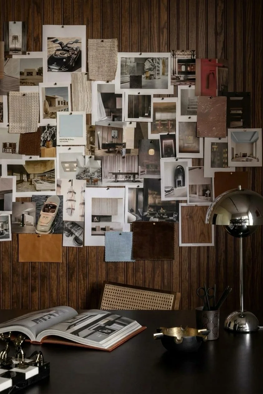 A wall covered with various interior design images and fabric swatches, a desk with an open magazine, a silver desk lamp, and a pen holder.