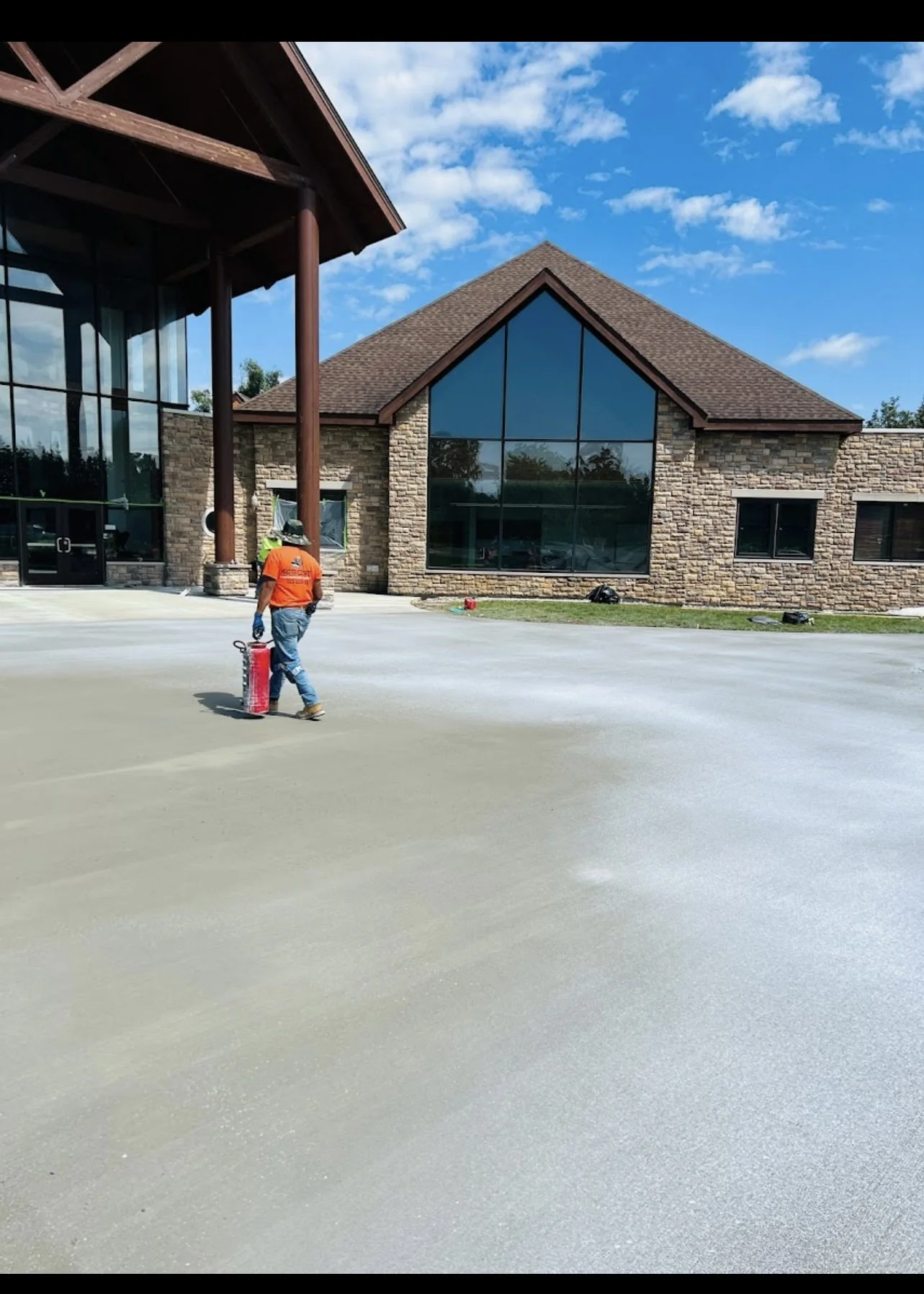 A worker wearing an orange shirt, jeans, and gloves walking on a freshly poured concrete surface outside a building with large glass windows and brick walls, under a blue sky with some clouds.