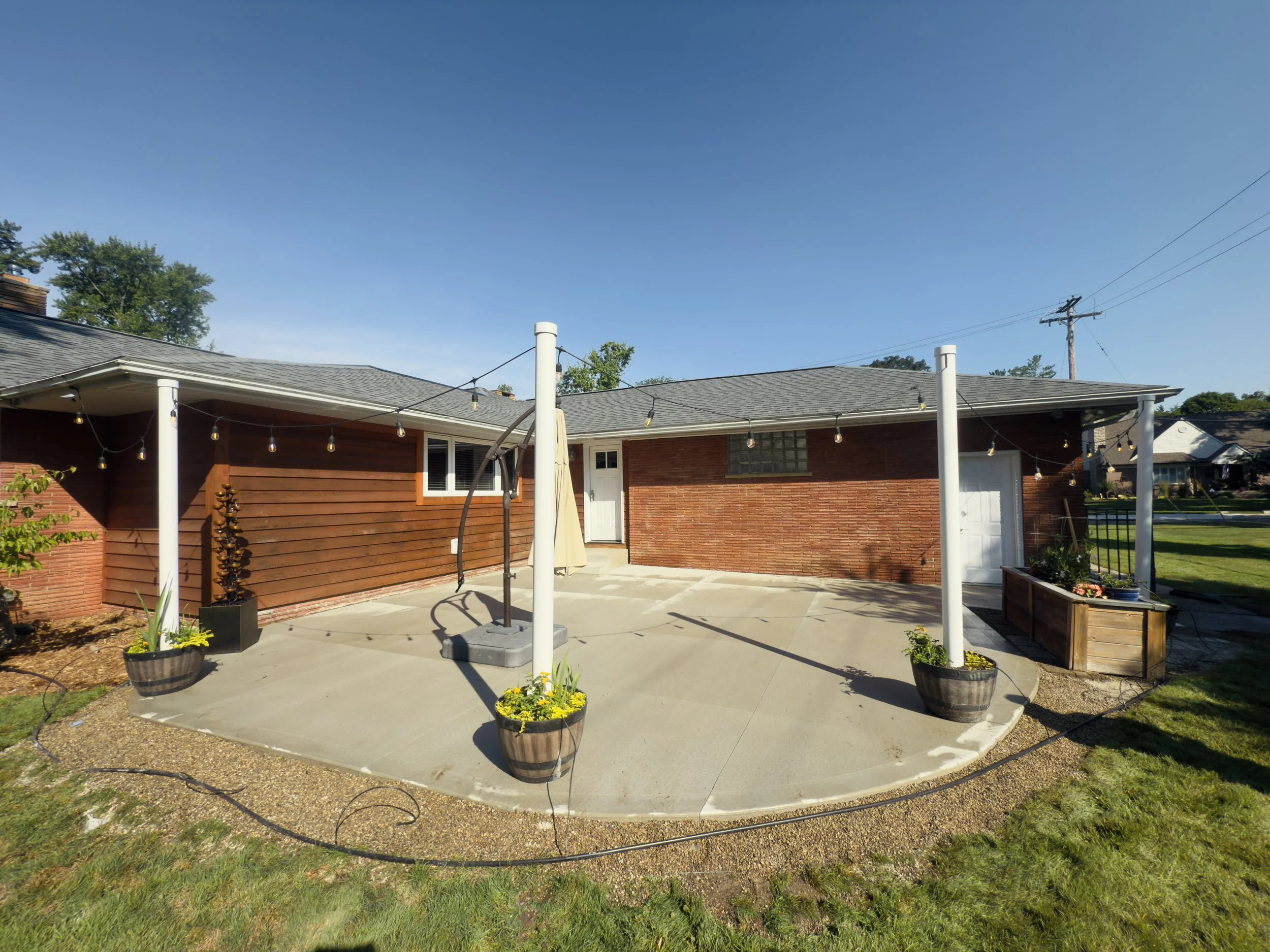Rear view of a house with a concrete patio, string lights, and planters with flowers, during sunny weather.