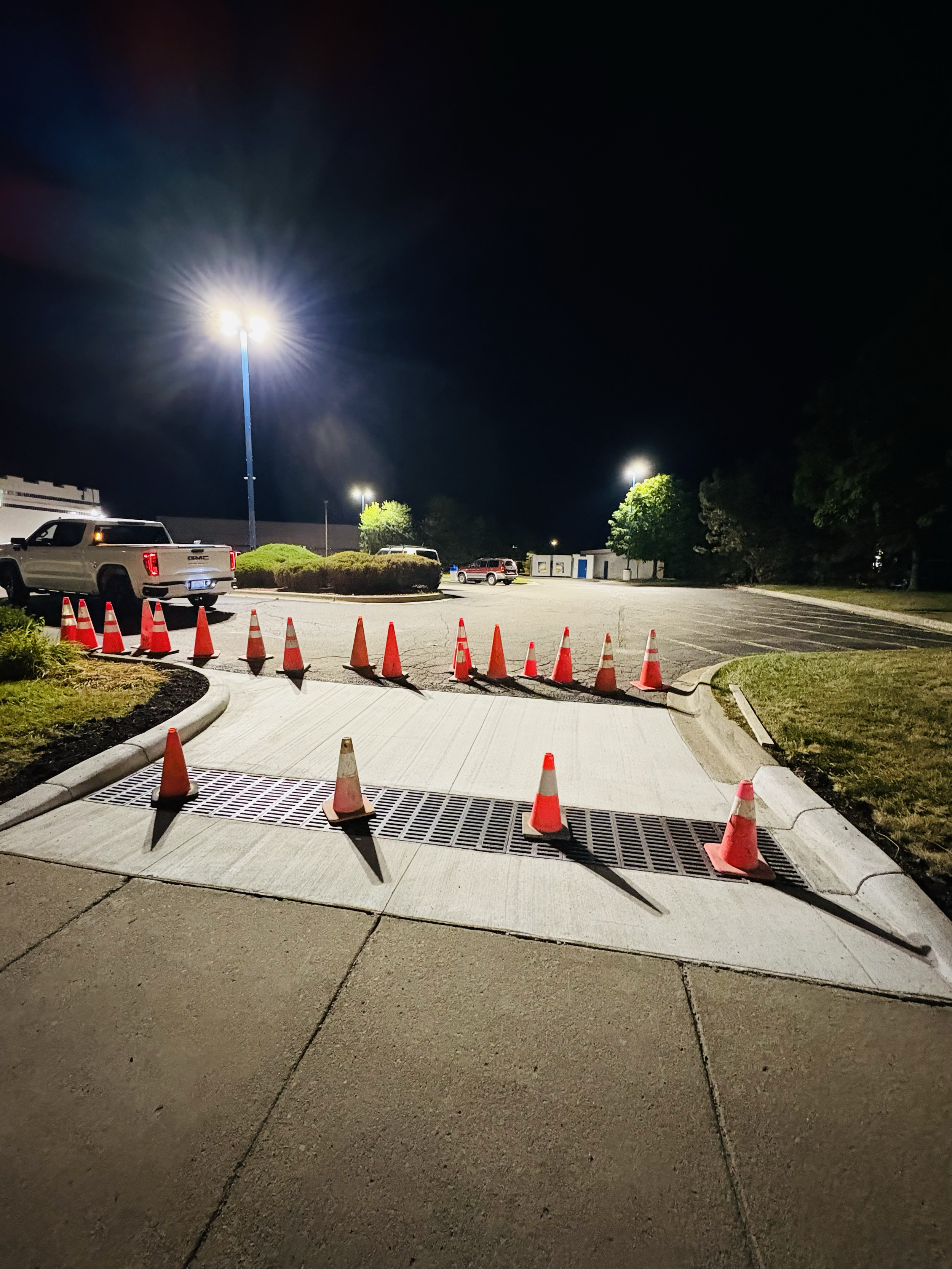 Nighttime view of a sidewalk with orange construction cones blocking the accessible ramp and drain grate, parking lot in the background, with streetlights illuminating the scene.