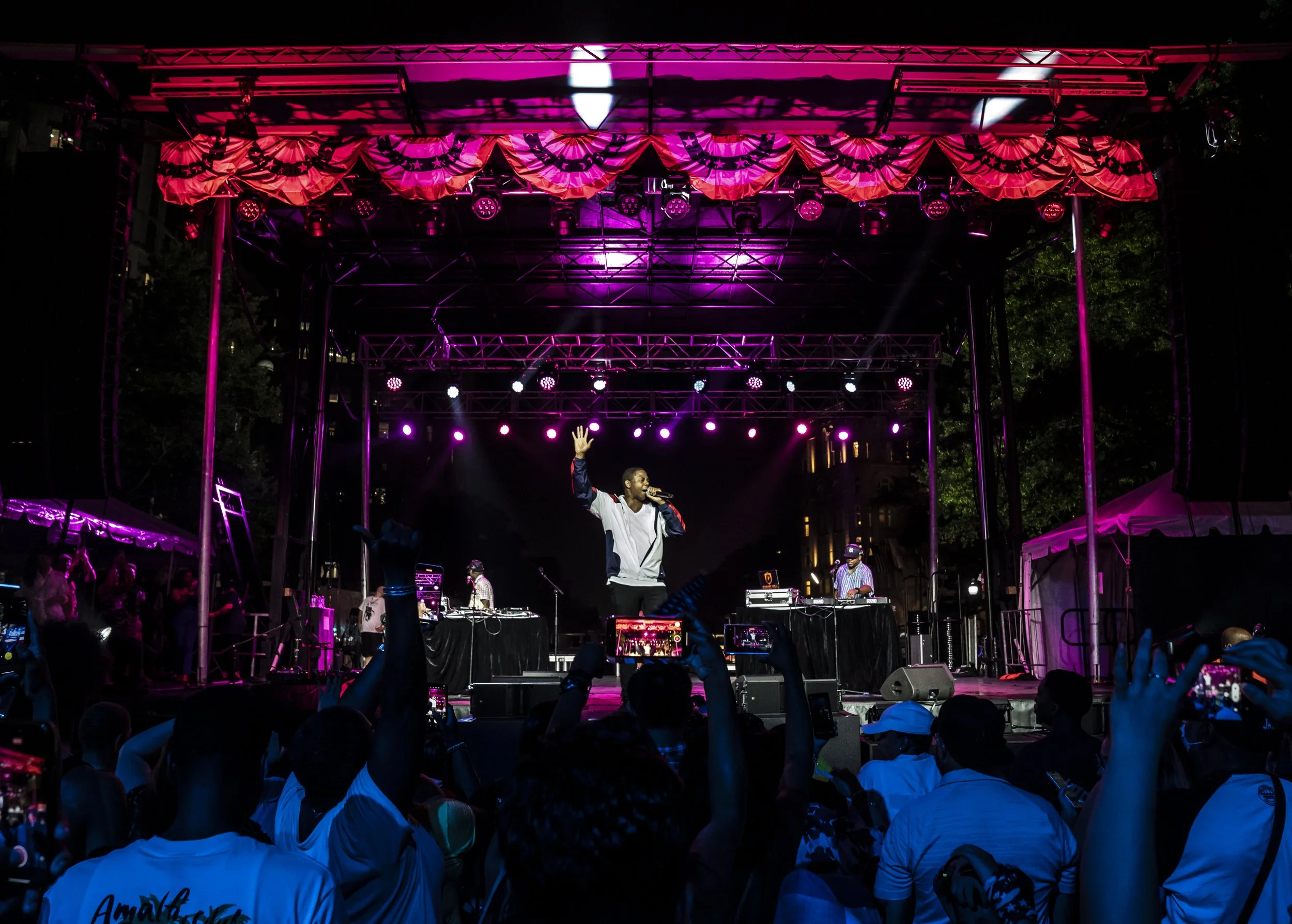 Nighttime outdoor concert performance with singer and DJ on stage lit with pink and purple lights, crowd in front watching.