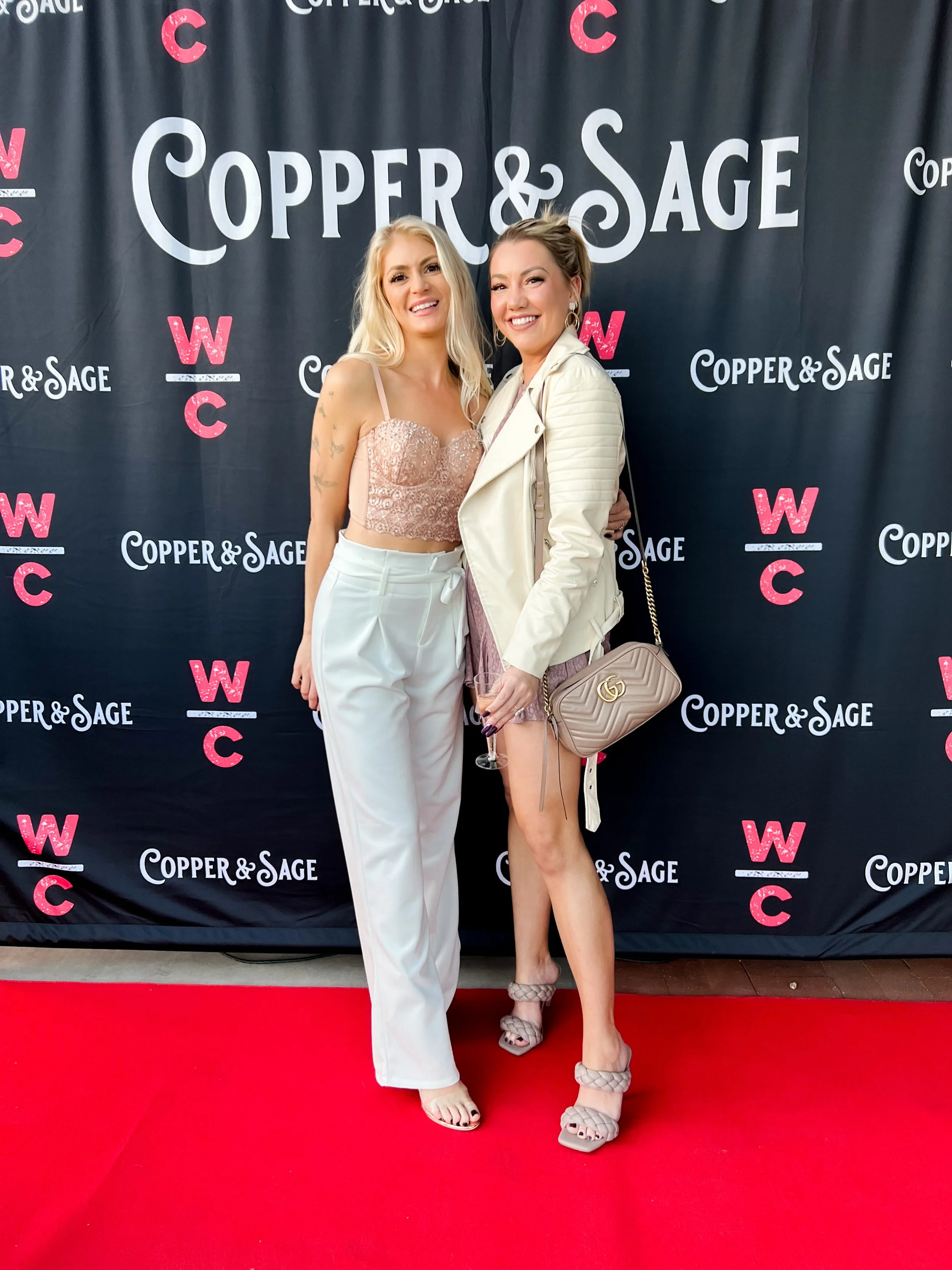 Two women standing on a red carpet in front of a black backdrop with the words 'Copper & Sage' and 'WC' repeatedly printed. The woman on the left has long blonde hair and is wearing a pink lace crop top and white high-waisted pants. The woman on the 