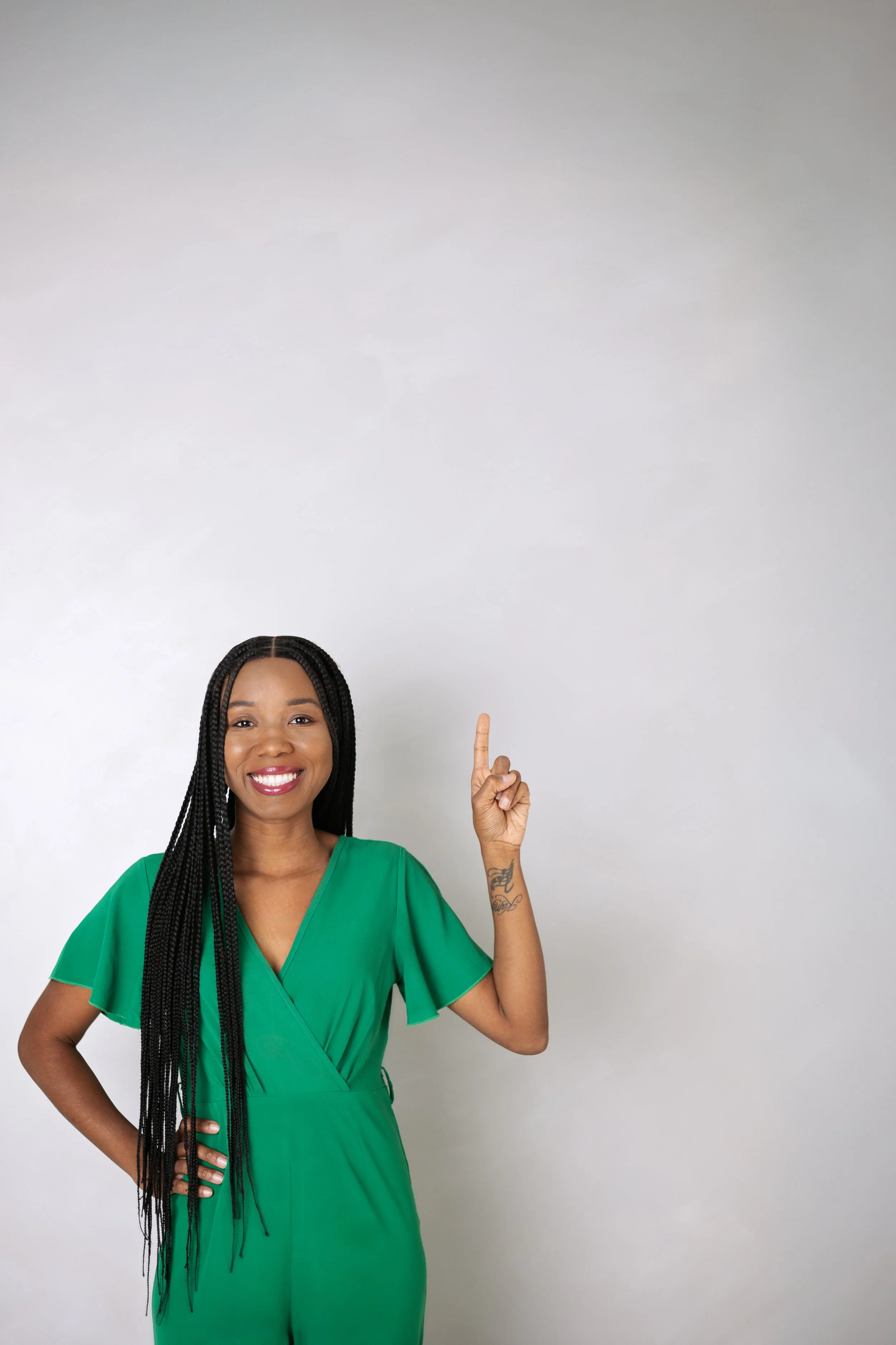 A woman wearing a green outfit smiling and pointing upward with her right index finger against a plain light grey background.
