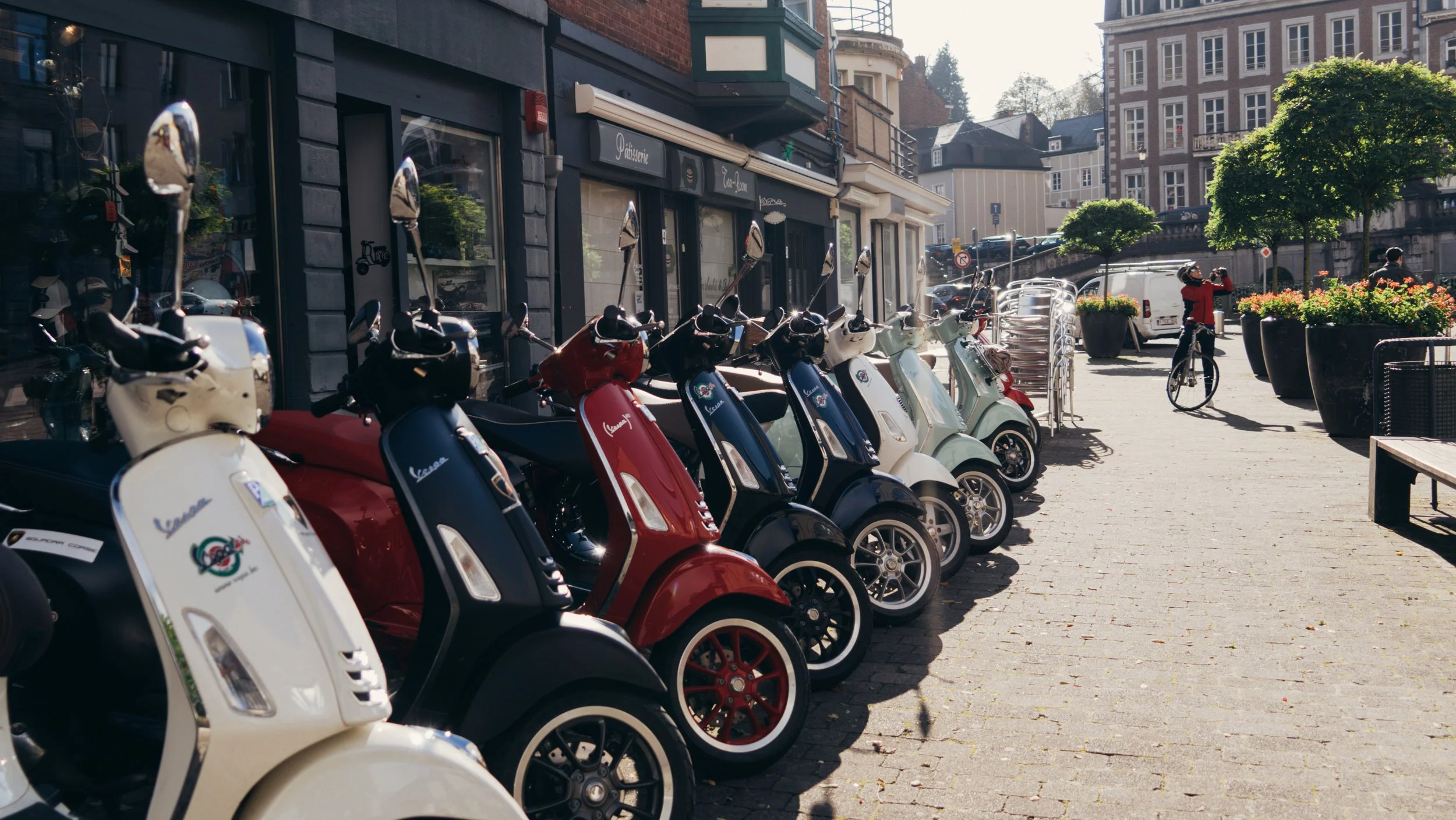 Row of scooters parked on a city sidewalk next to a storefront, with a person riding a bicycle in the background under trees and buildings.