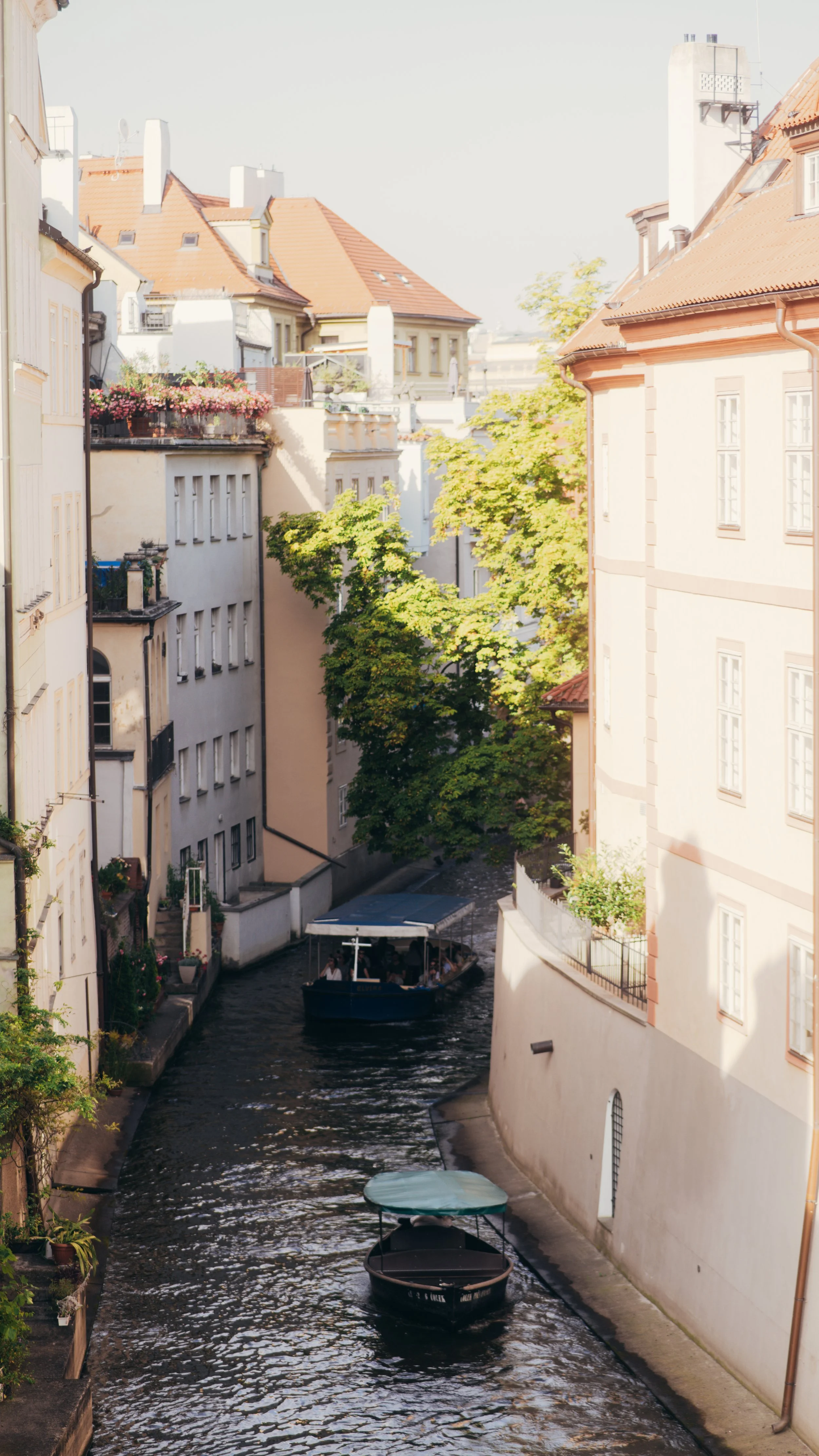 A narrow canal in a European city with boats, surrounded by tall, light-colored buildings and trees.