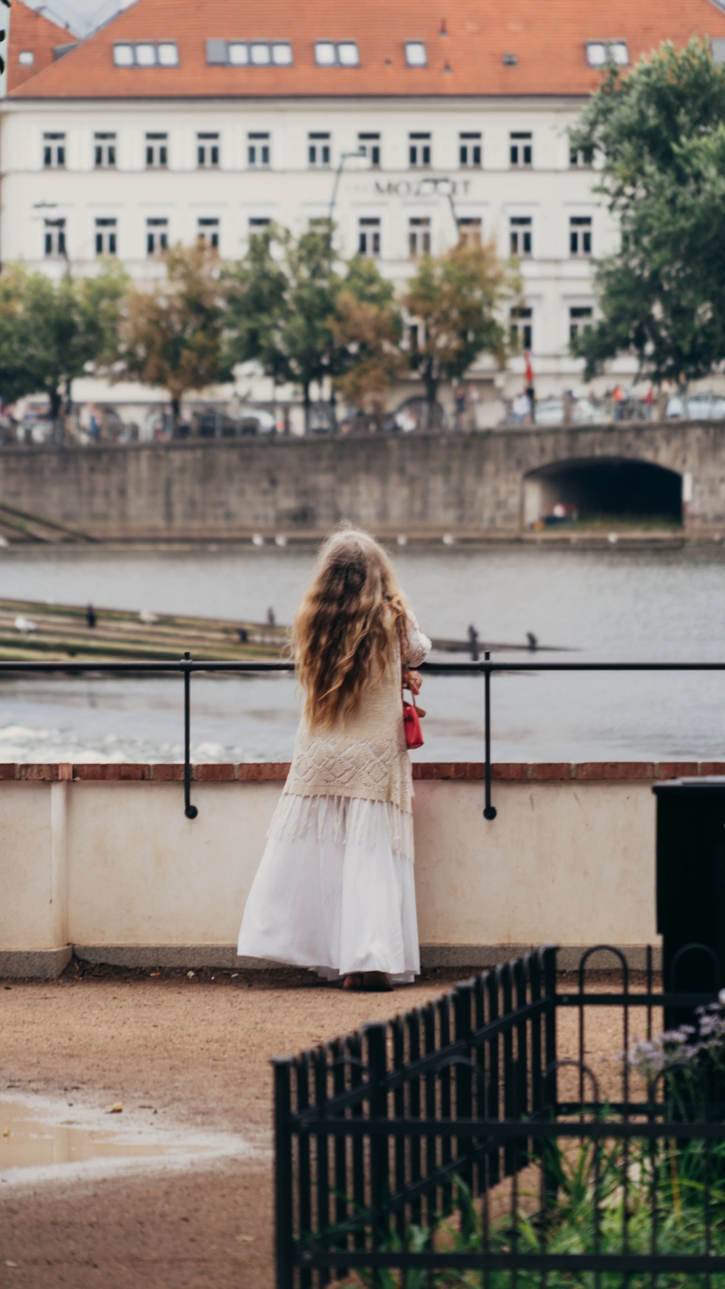 A young girl with long, wavy hair wearing a white dress and a beige knitted shawl standing by a canal.