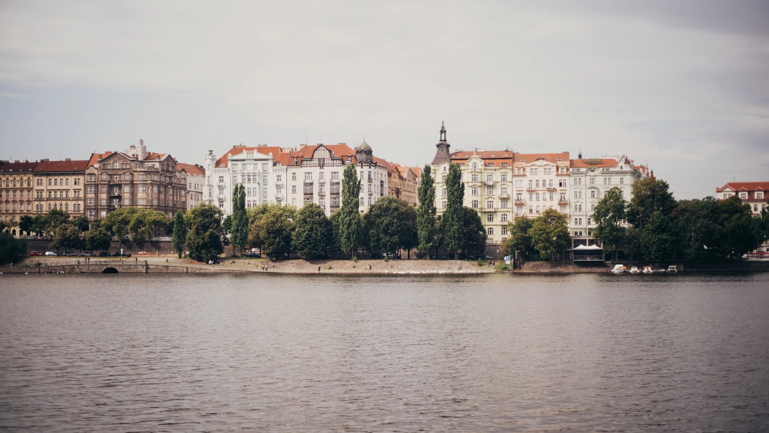 View of a riverside cityscape with colorful historic buildings, trees lining the riverbank, and several small boats docked by the shore on a cloudy day.