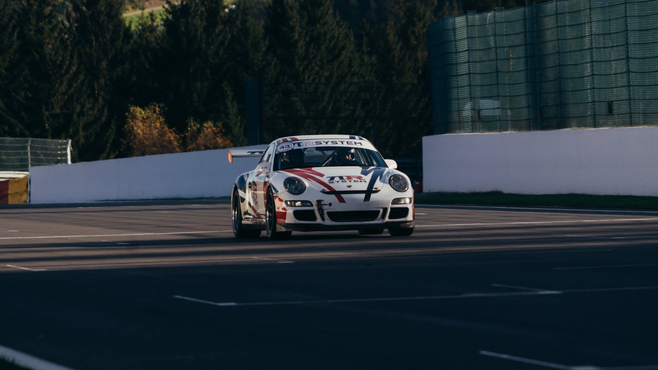 A white race car with red and black decals on a race track, with trees and a barrier wall in the background.