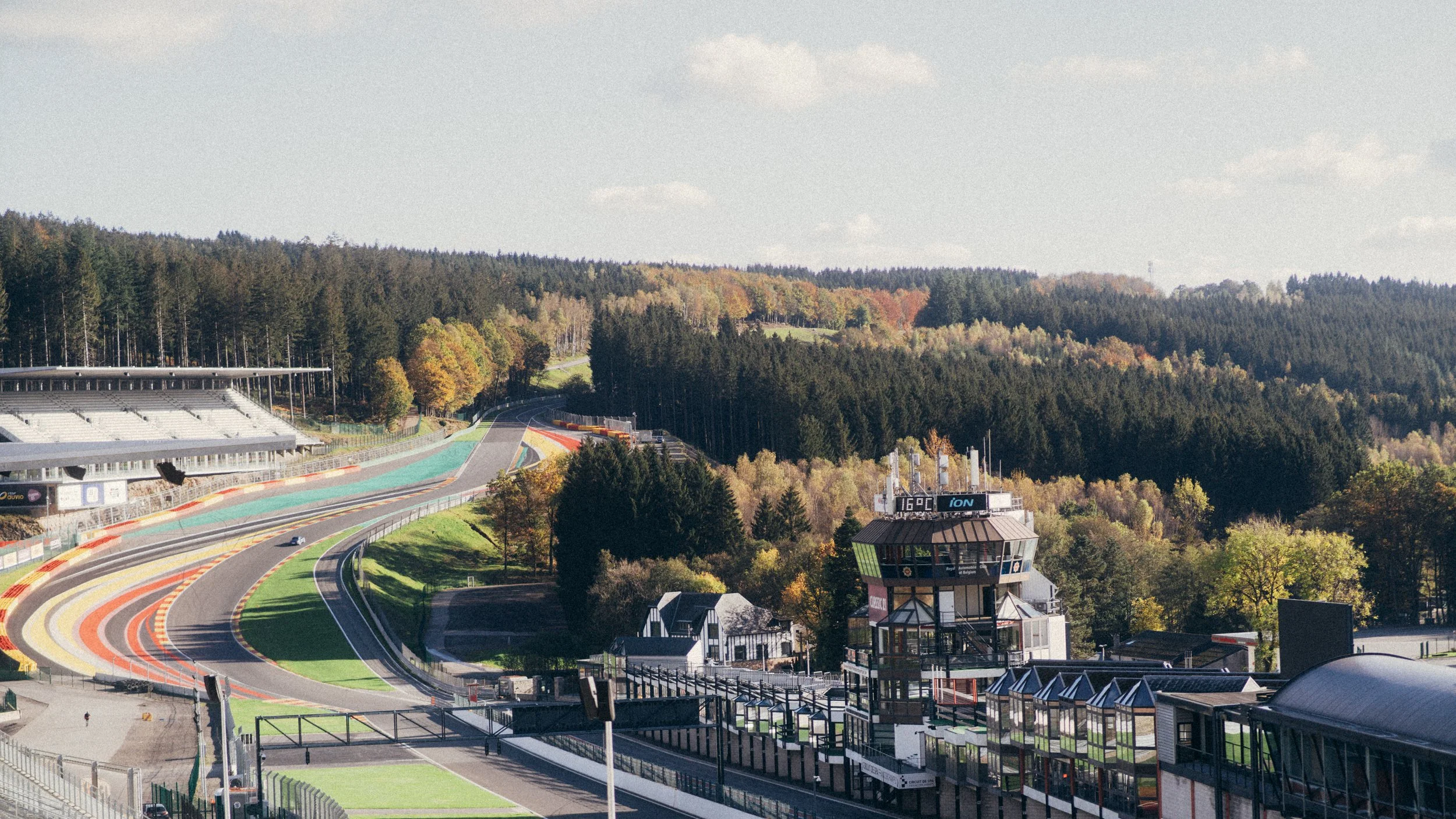 A race circuit with empty grandstands, surrounded by a forested hillside with fall foliage, featuring a control tower and paved viewing areas.
