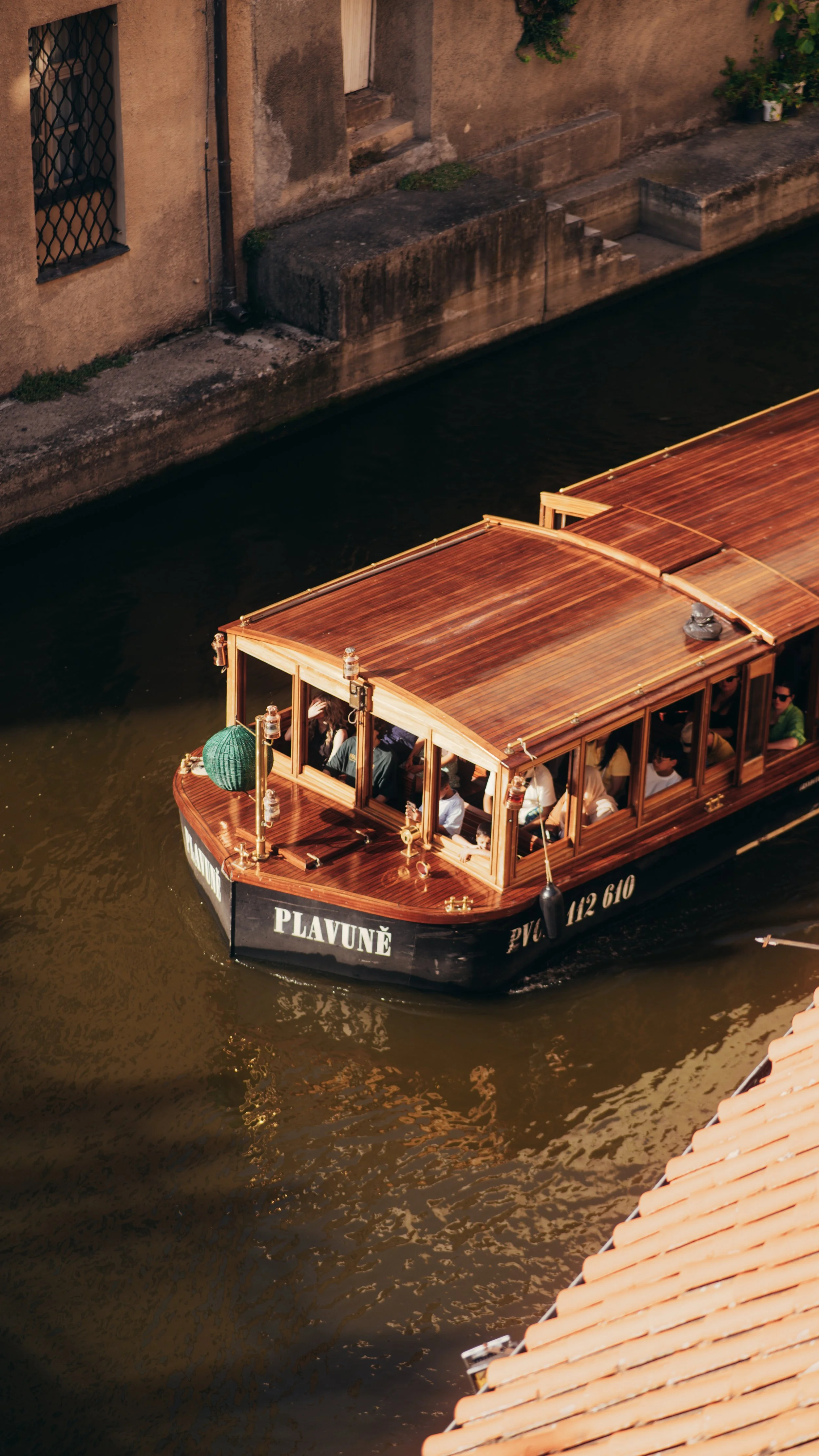A wooden boat named 'PLAVUŇĖ' is on a narrow waterway, carrying passengers through a city with old buildings and tiled rooftops.