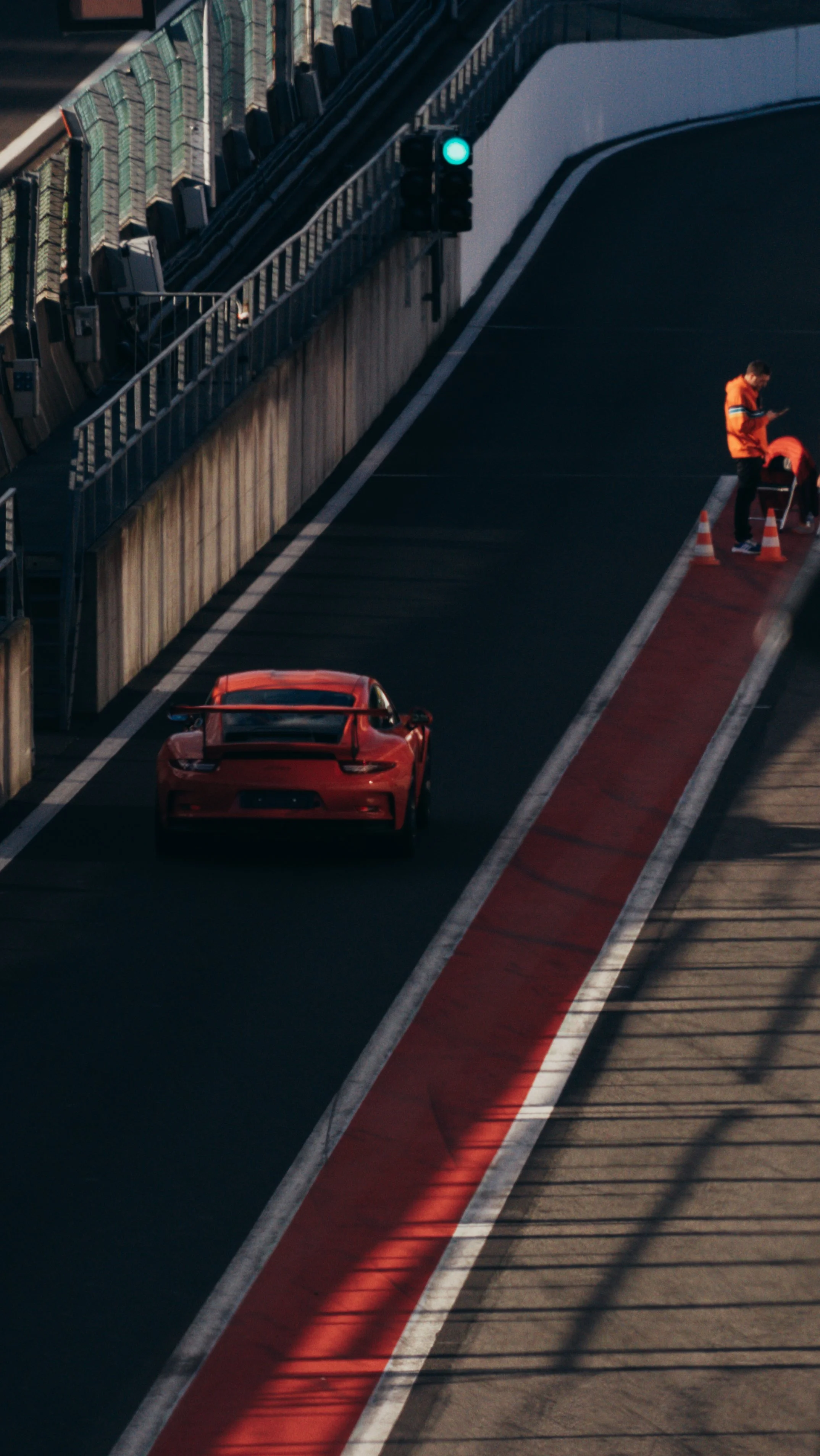 A red sports car on a racetrack with two staff members in orange jackets next to it, one working with traffic cones and the other looking at a phone, under a green traffic light.