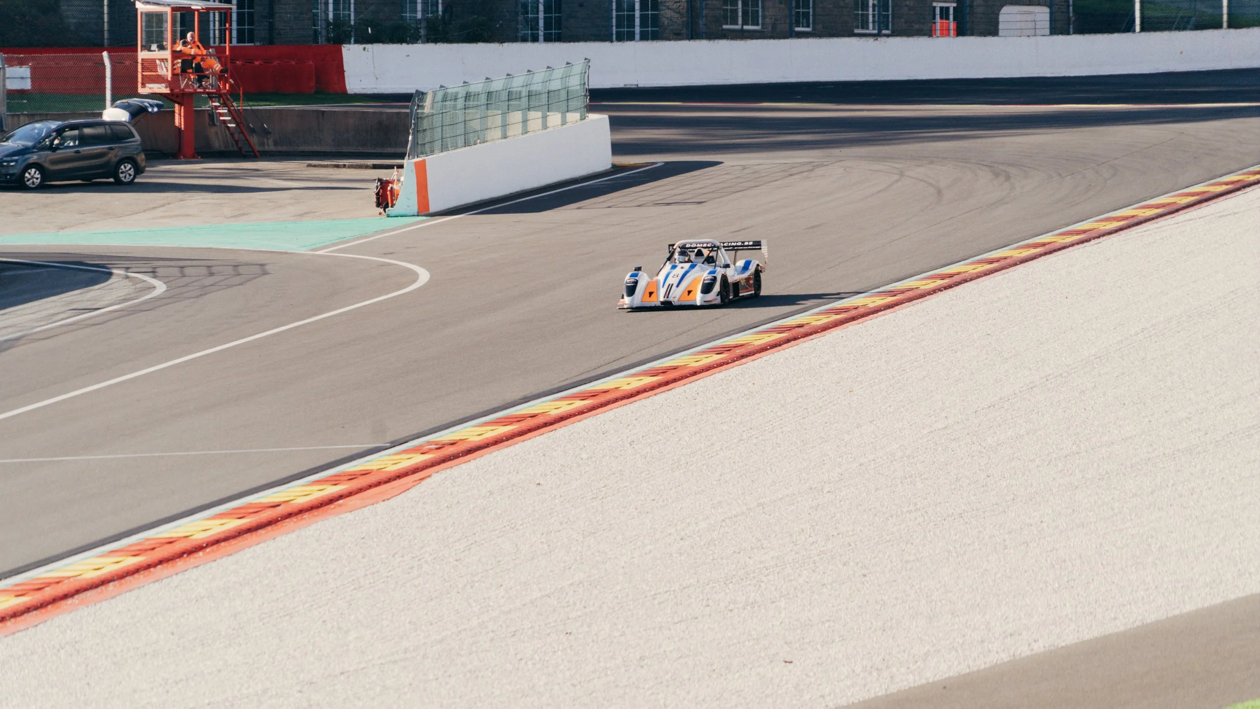 A race car driving on a racing track with orange, yellow, and red curbs, with a red safety station in the background.