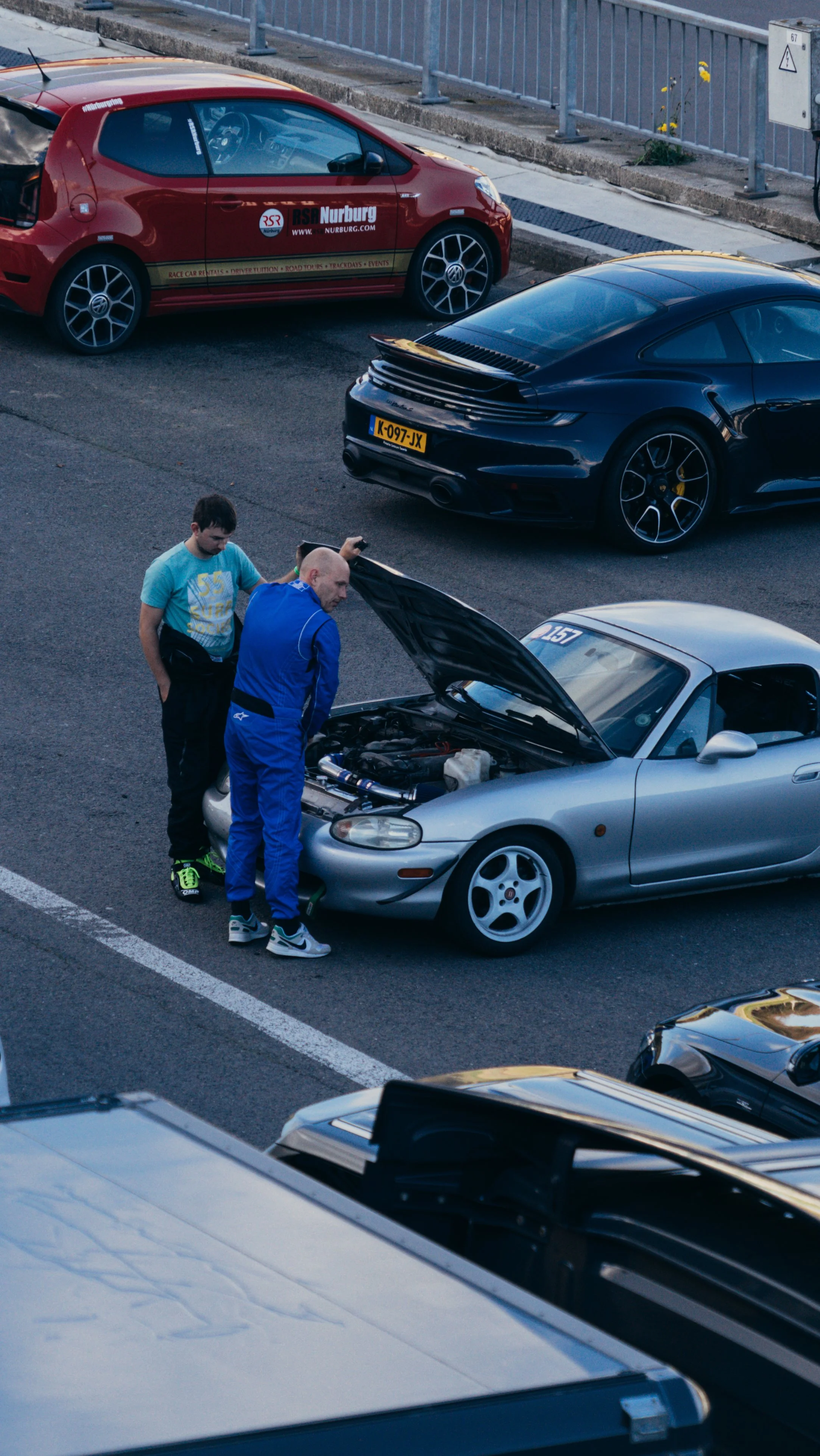Two men are checking the engine of a silver sports car with its hood open in a parking lot. Other cars are parked nearby.