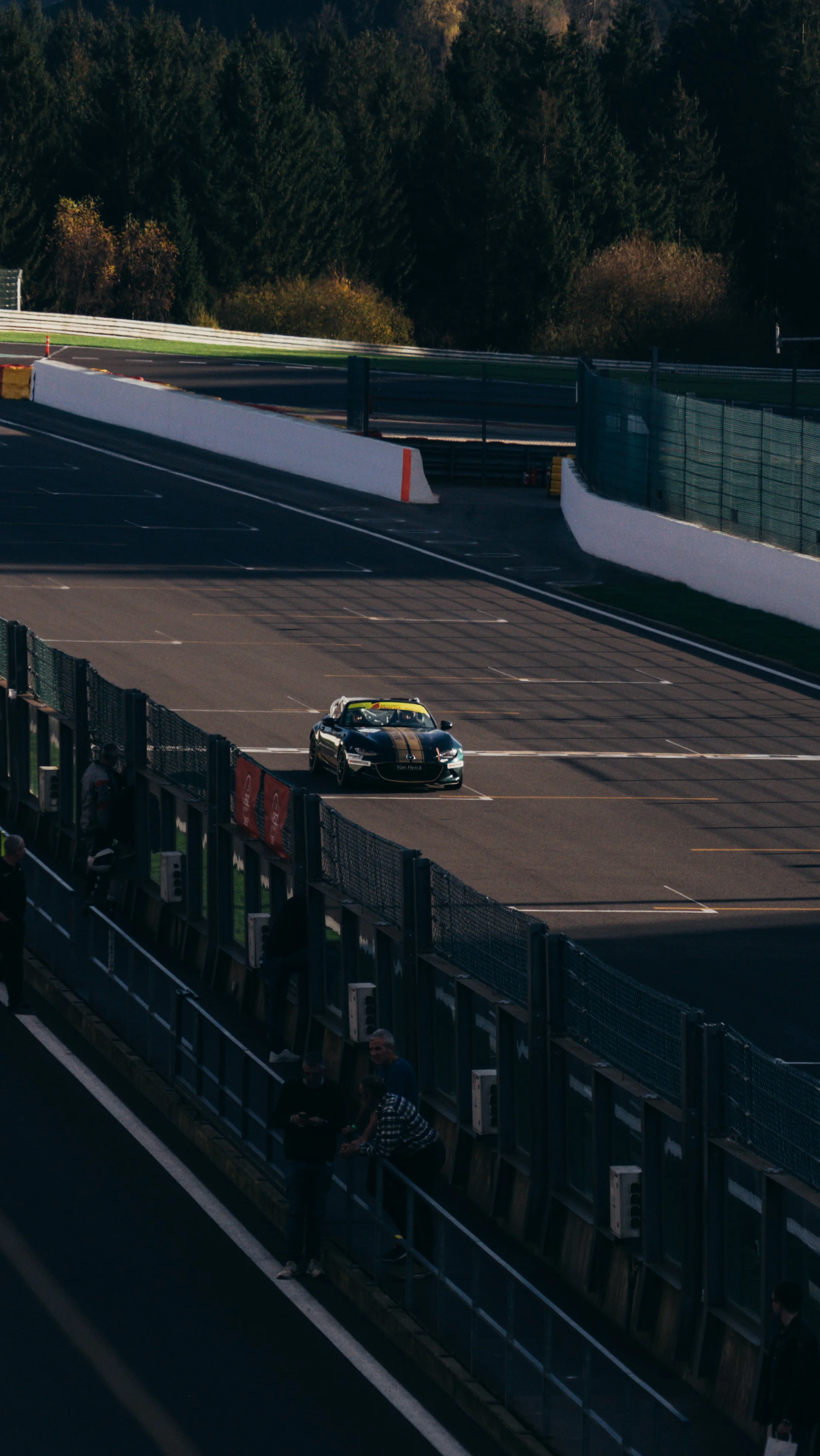 A race car on a race track during the day, with spectators standing behind a barrier.