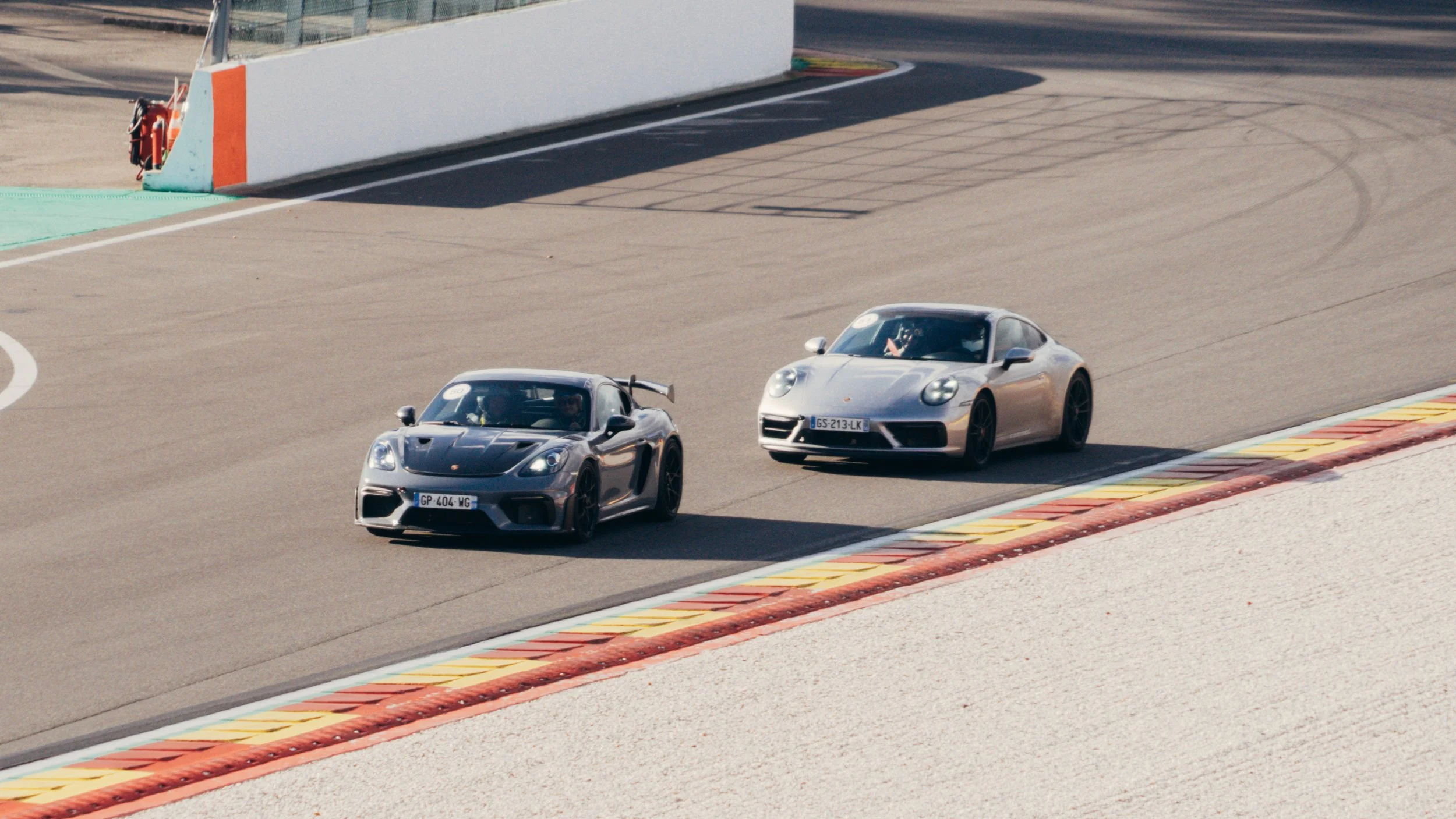Two sports cars, one black and one silver, racing on a track with curbing. The black car is in front, and both are driving on a curved section of the race track with a barrier on the left side.