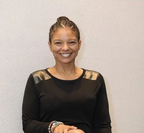 Woman smiling, wearing a black top with leather details on shoulders, standing against a textured white wall.