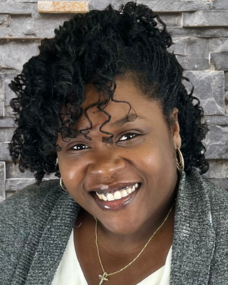 A woman with dark curly hair, smiling, wearing hoop earrings and a necklace, against a stone wall background.