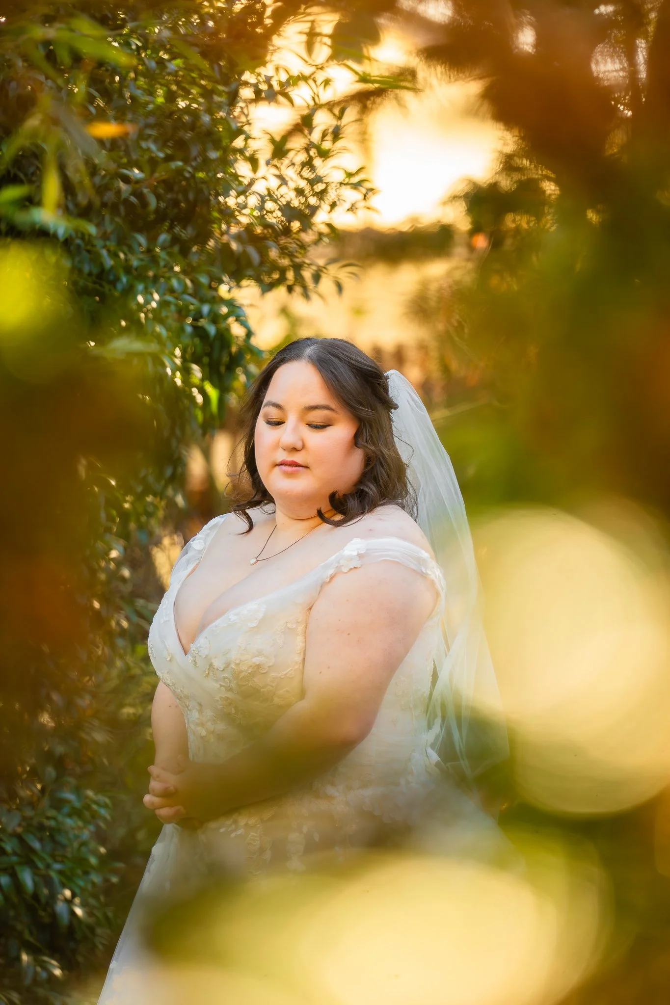 A bride in a wedding dress standing outdoors during sunset, surrounded by blurred greenery and warm bokeh lights.
