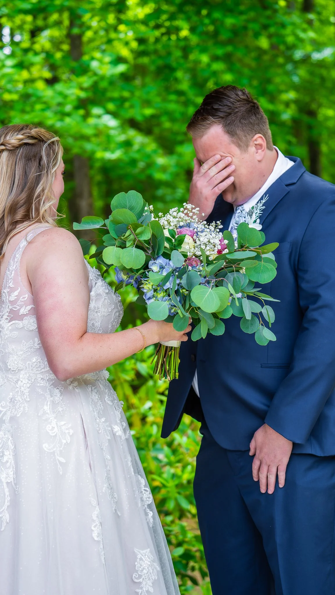 A bride holding a bouquet of flowers and a groom covering his face with his hand during a wedding ceremony outdoors.