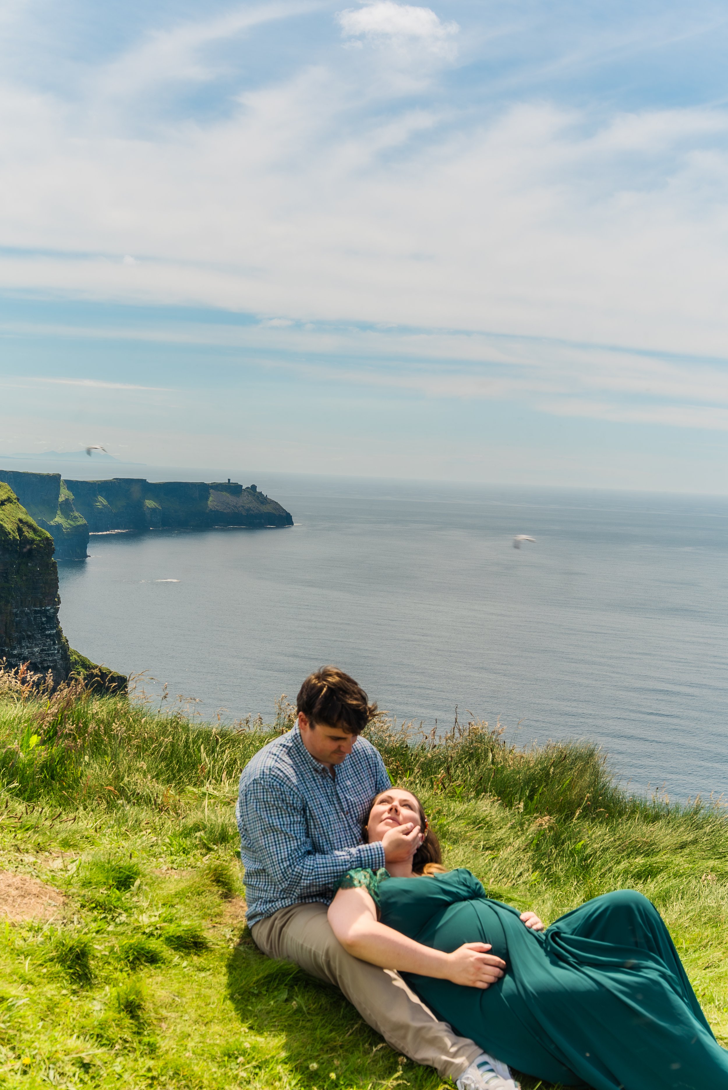 A man and woman on a grassy hilltop near the ocean, with cliffs and a cloudy sky in the background. The woman is lying down, and the man is holding her chin, leaning over her.