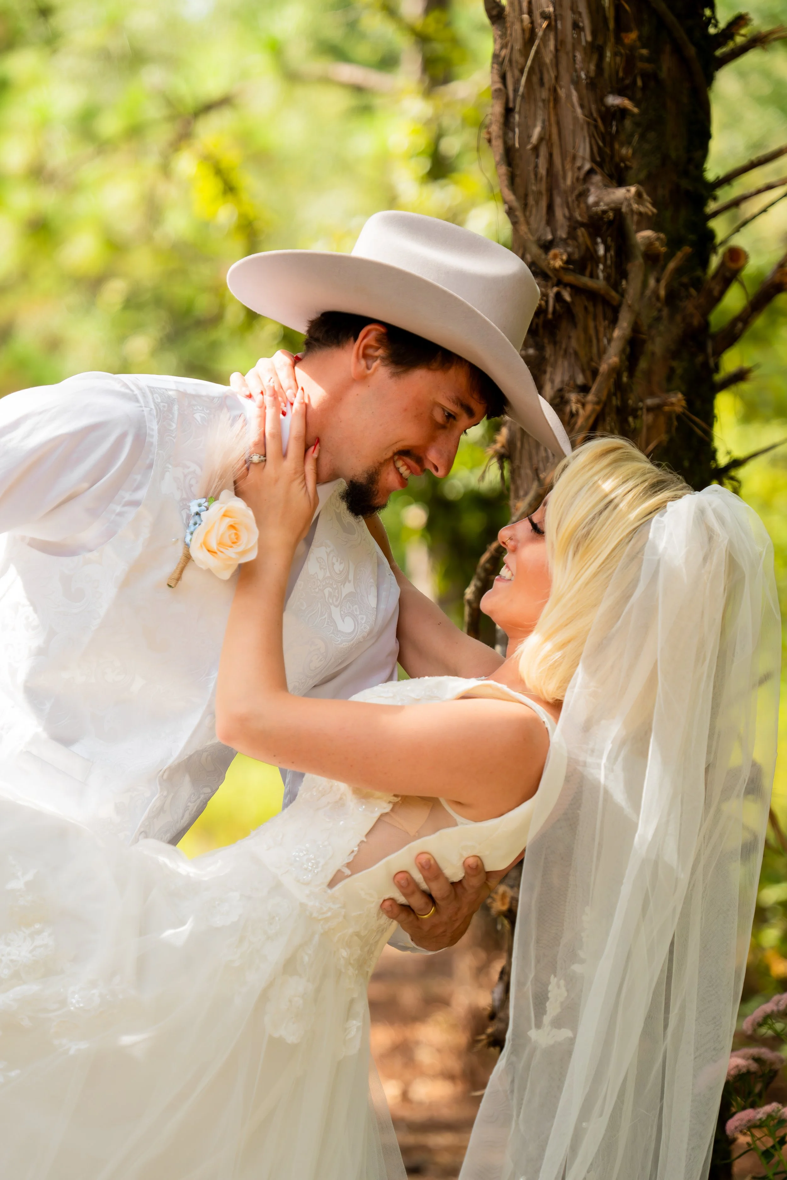 A joyful bride and groom in wedding attire, smiling and looking at each other affectionately in an outdoor setting with trees.