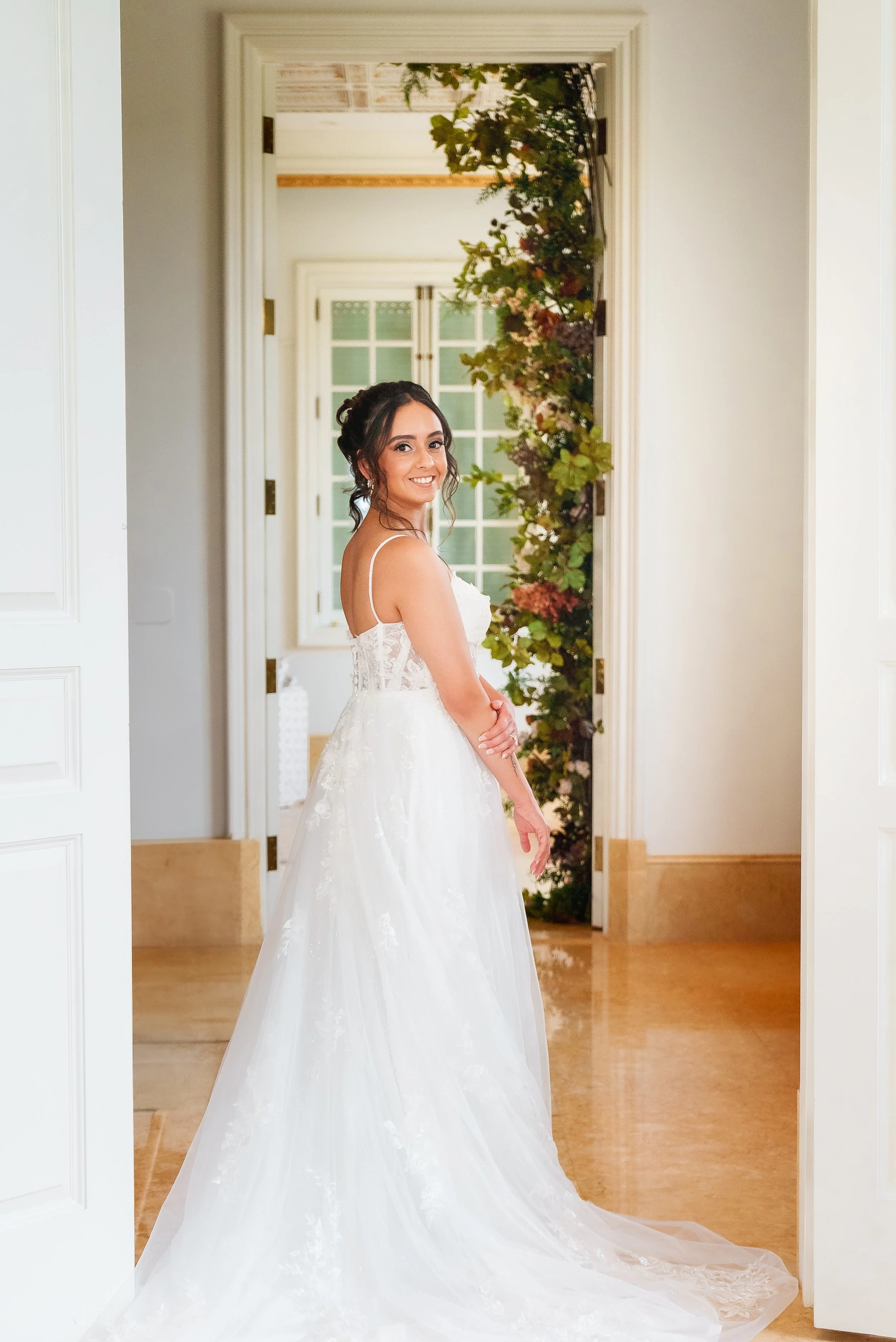 A bride in a white wedding dress standing in a doorway, smiling, with flowers and greenery outdoor in the background.