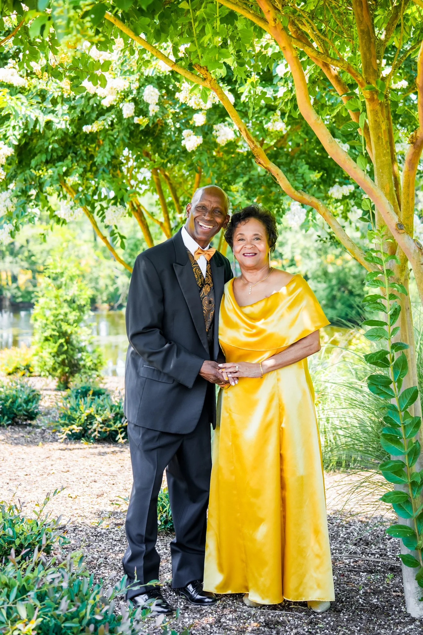 An elderly couple dressed in formal attire, standing outdoors under a green tree with white flowers, smiling and holding hands near a body of water.