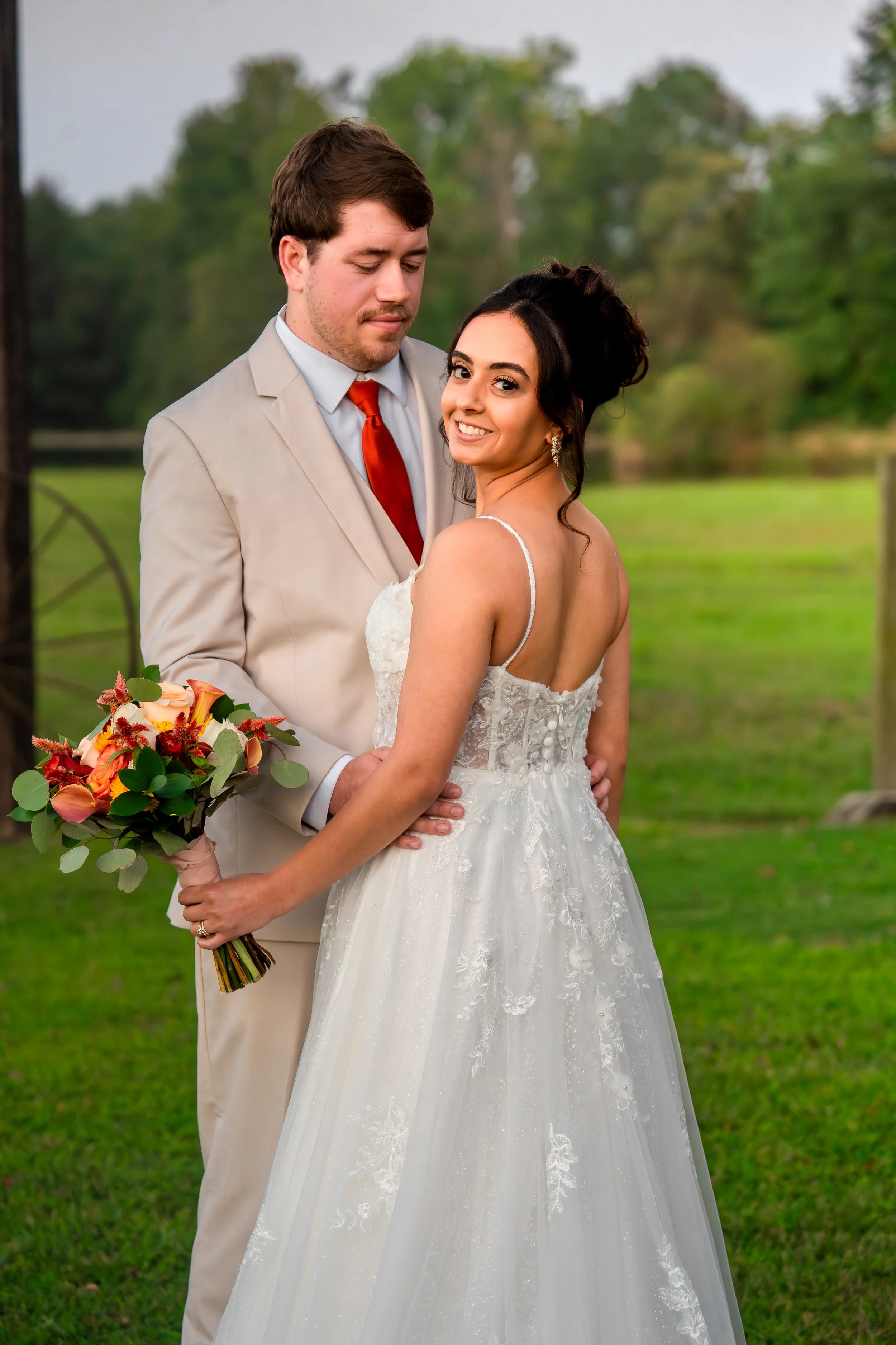Bride and groom standing outdoors on their wedding day, with the groom holding a bouquet and the bride smiling at the camera. The bride wears a white wedding dress with lace details, and the groom is in a light beige suit with a red tie. Green trees
