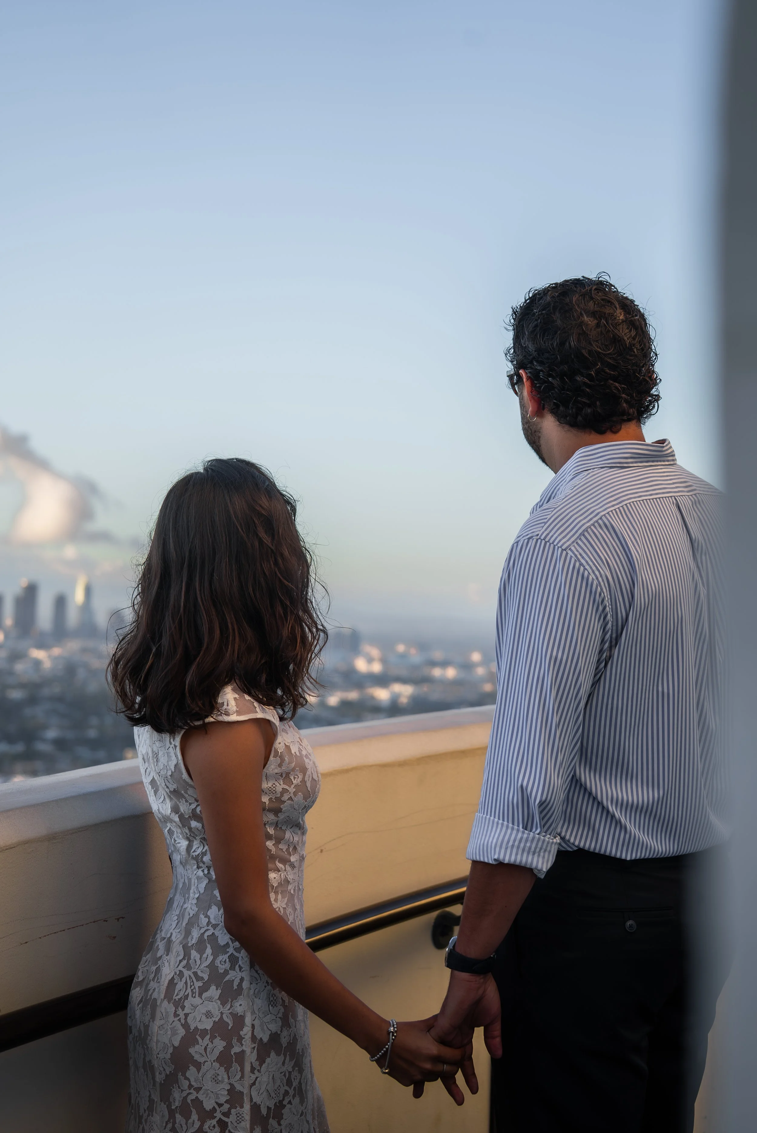 A man and woman are holding hands, facing each other on a balcony overlooking a city skyline with a large building and a hot air balloon in the sky.
