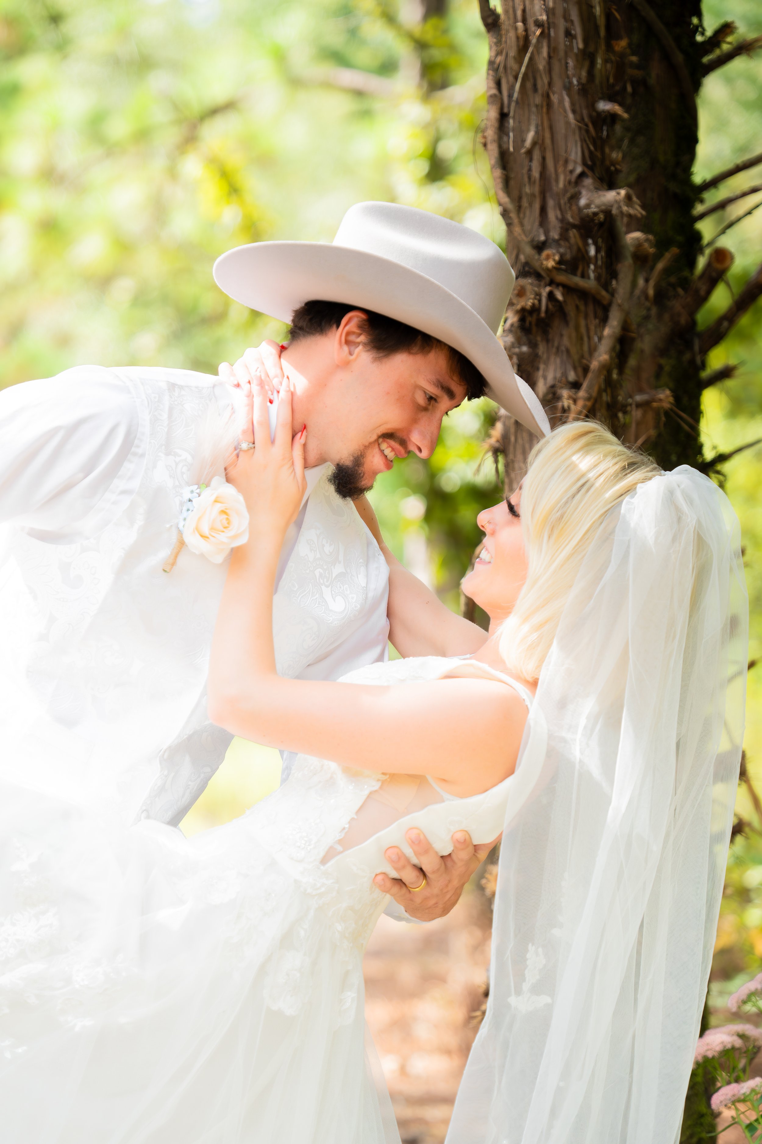 A groom lifting a bride in a white wedding dress outdoors, both smiling and gazing at each other.