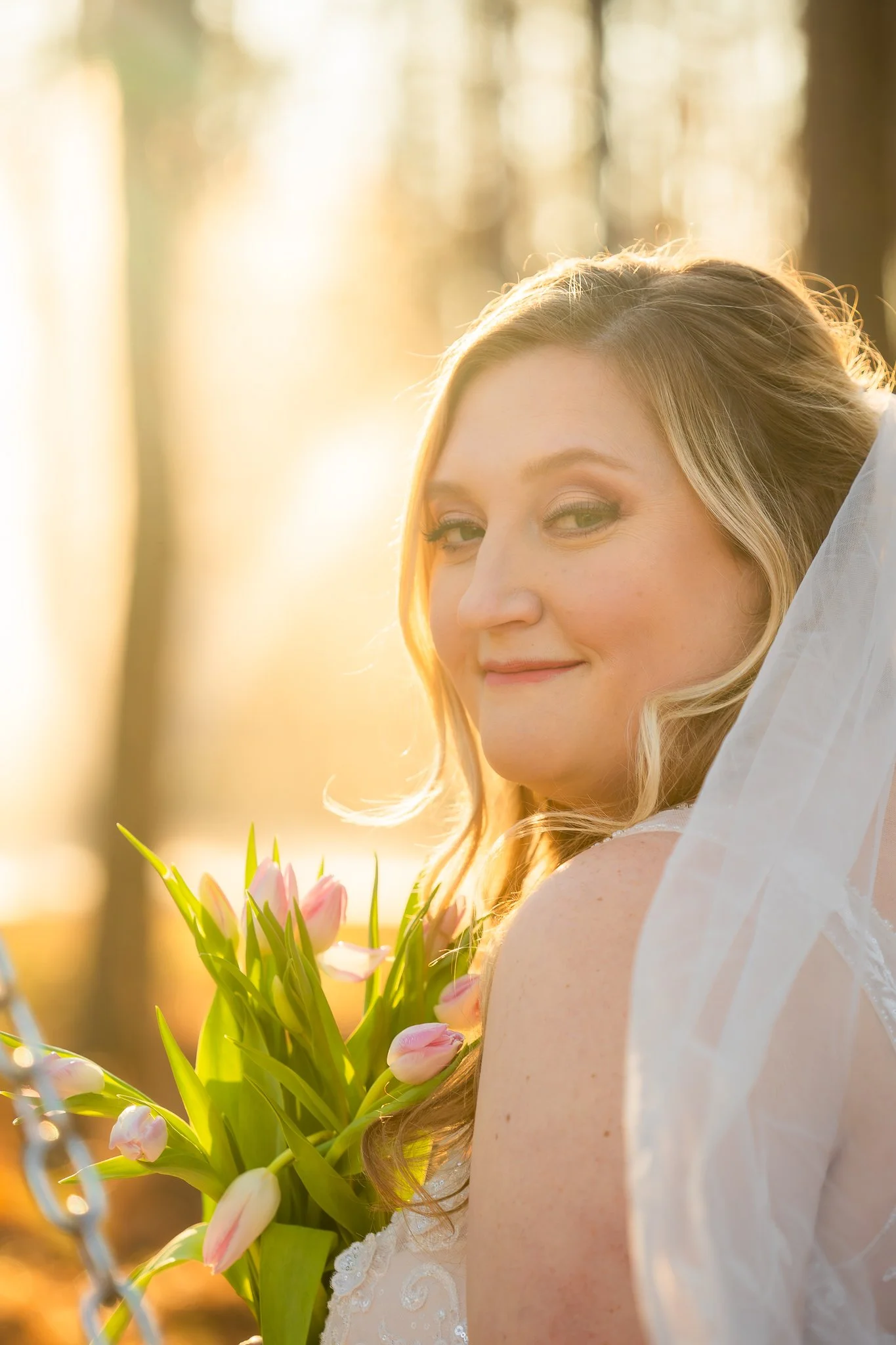 A woman in a wedding dress holding a bouquet of pink and white tulips, with sunlight streaming through trees behind her.