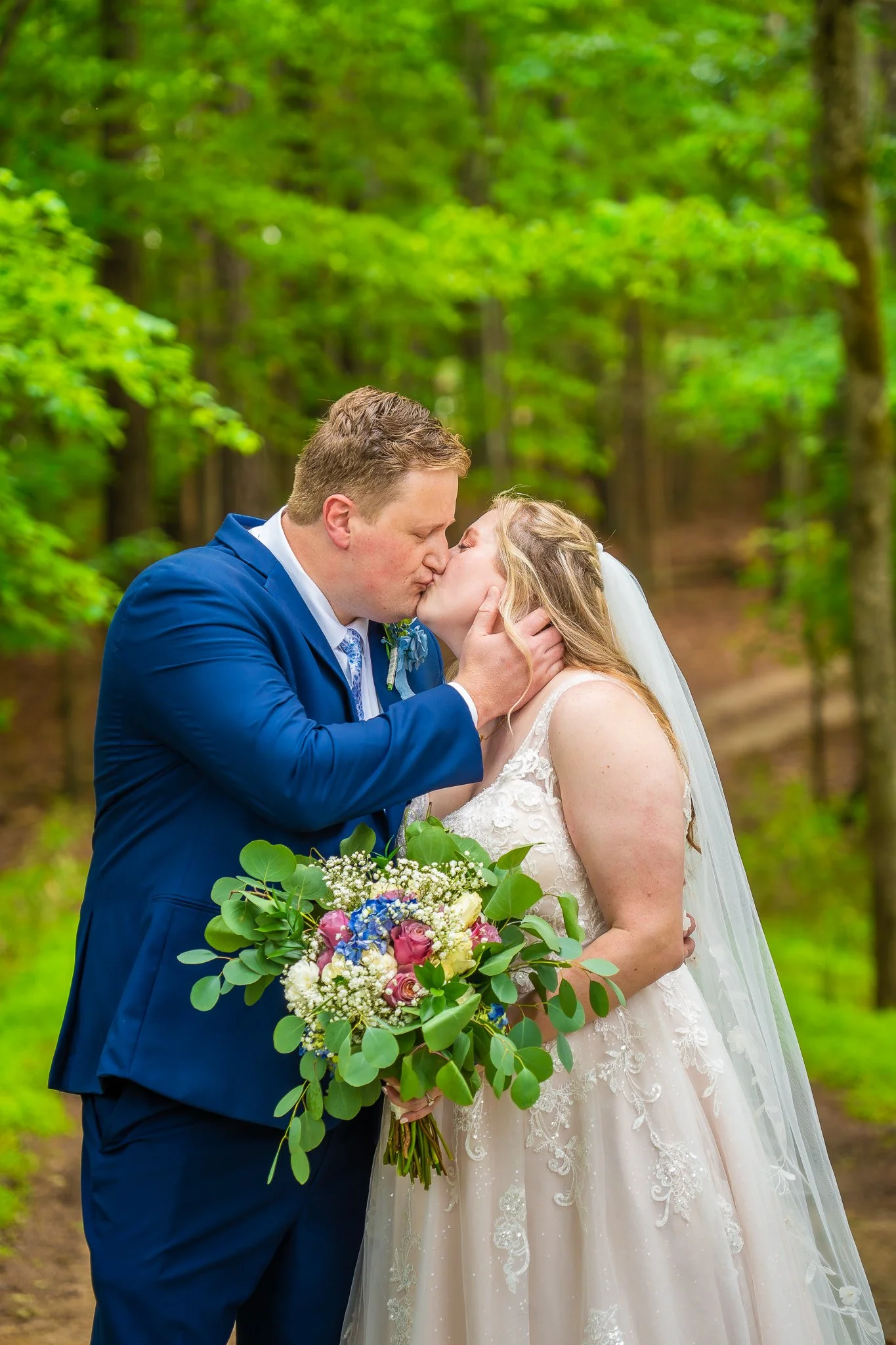A newlywed couple sharing a kiss in a green, wooded outdoor setting. The bride is holding a bouquet of pink, purple, white, and blue flowers with green foliage, while wearing a lace wedding dress and veil. The groom is dressed in a blue suit with a t
