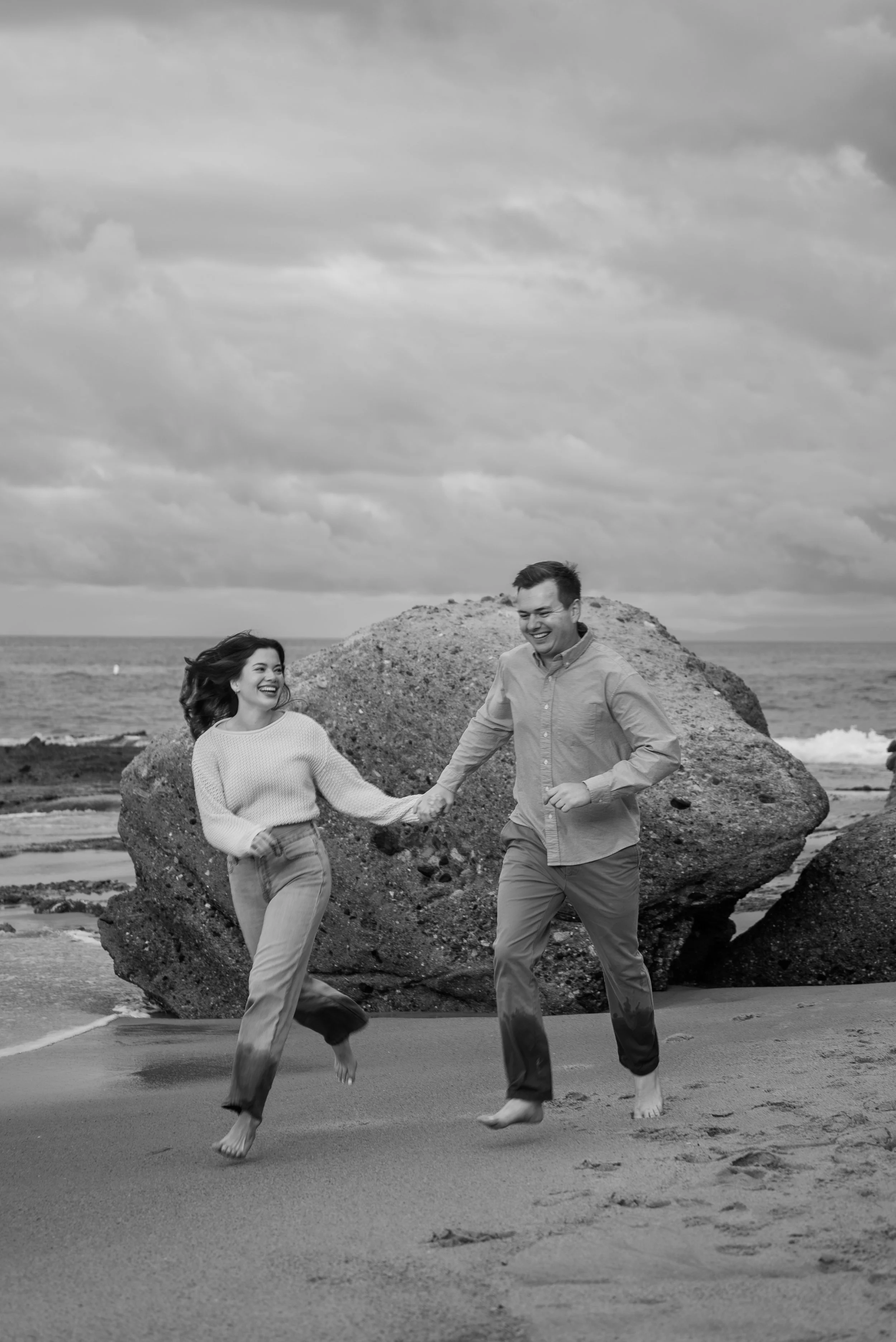A black-and-white photo of a man and woman holding hands and running barefoot on a beach with large rocks and ocean in the background, under a cloudy sky.