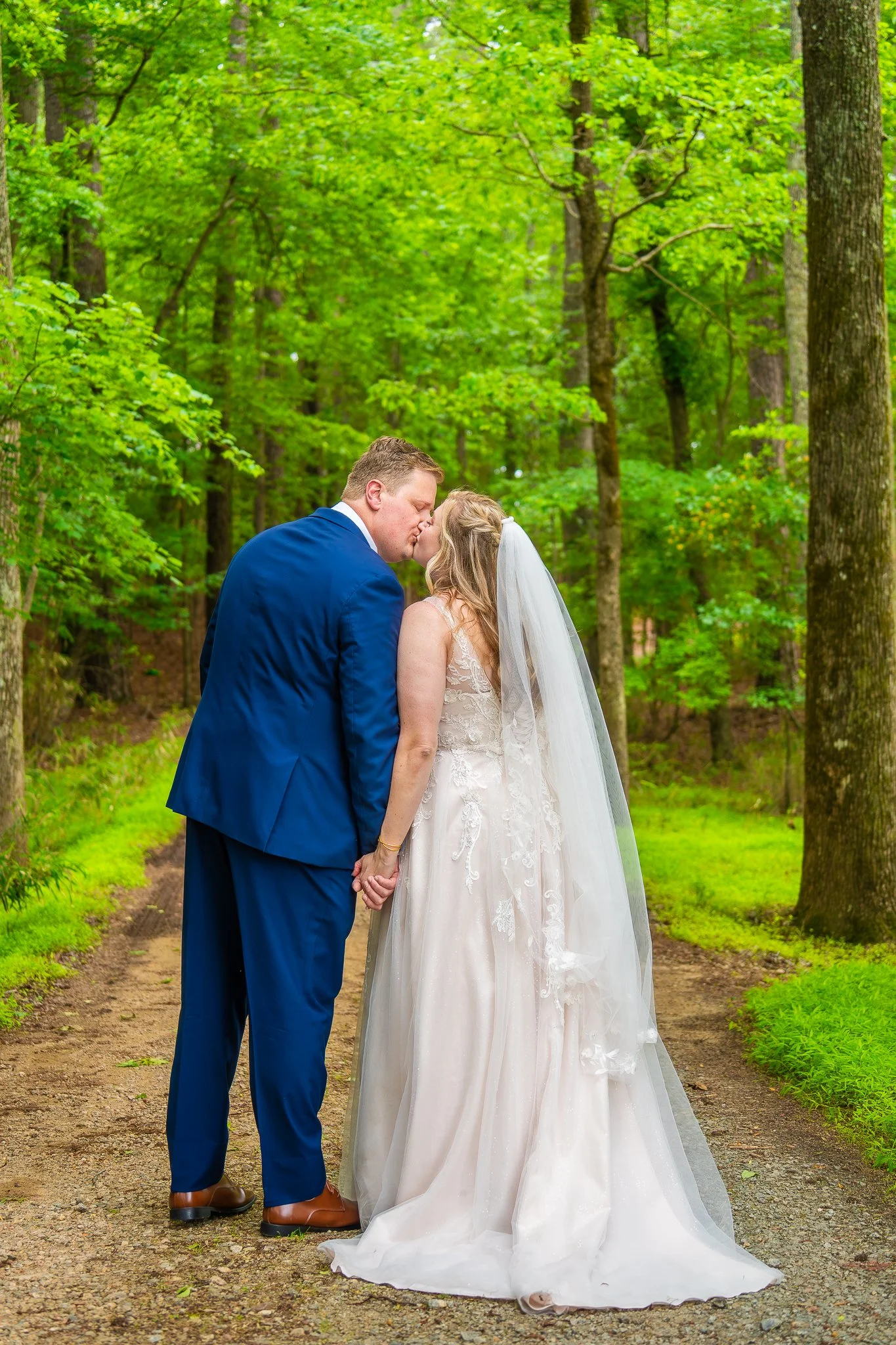 A bride and groom sharing a kiss on a forest path surrounded by green trees.