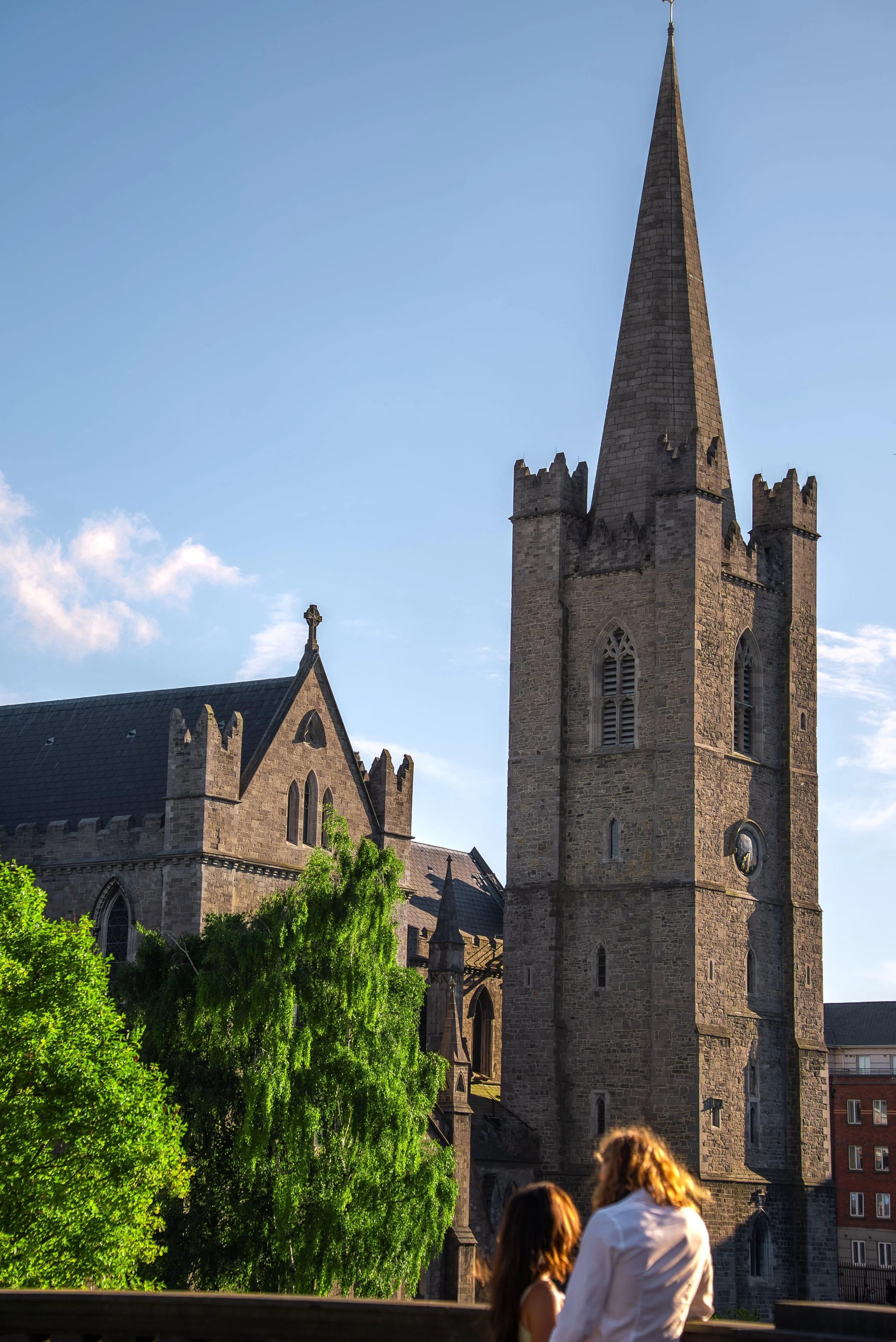 A church with a tall spire and Gothic architecture, with trees and people in the foreground, and a clear blue sky in the background.