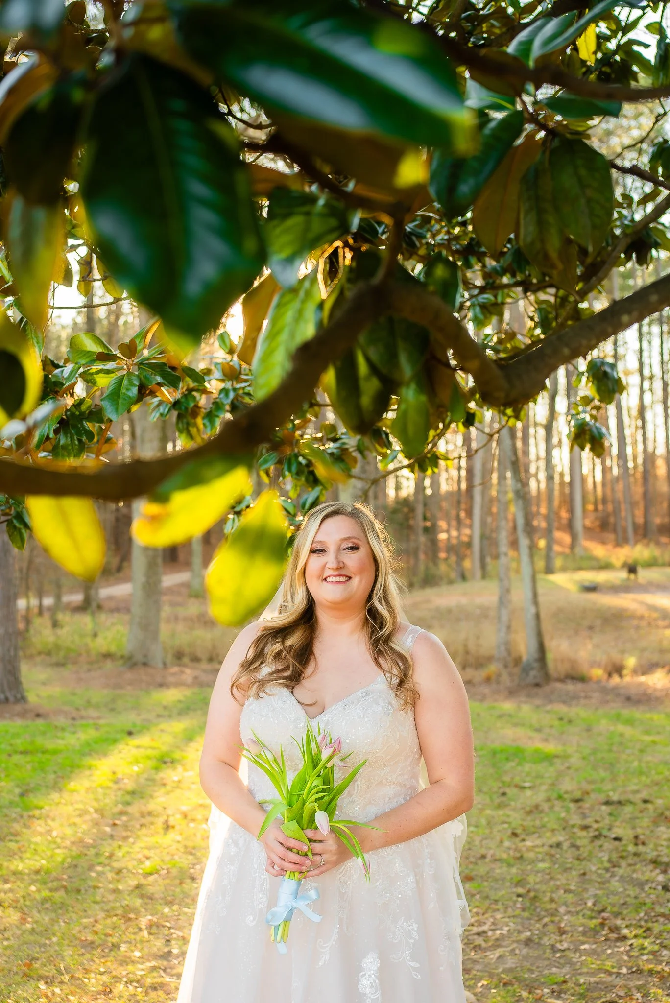 A bride in a white wedding dress holding a small bouquet of pink tulips outdoors during sunset, smiling at the camera, with trees in the background and sunlight filtering through the leaves.