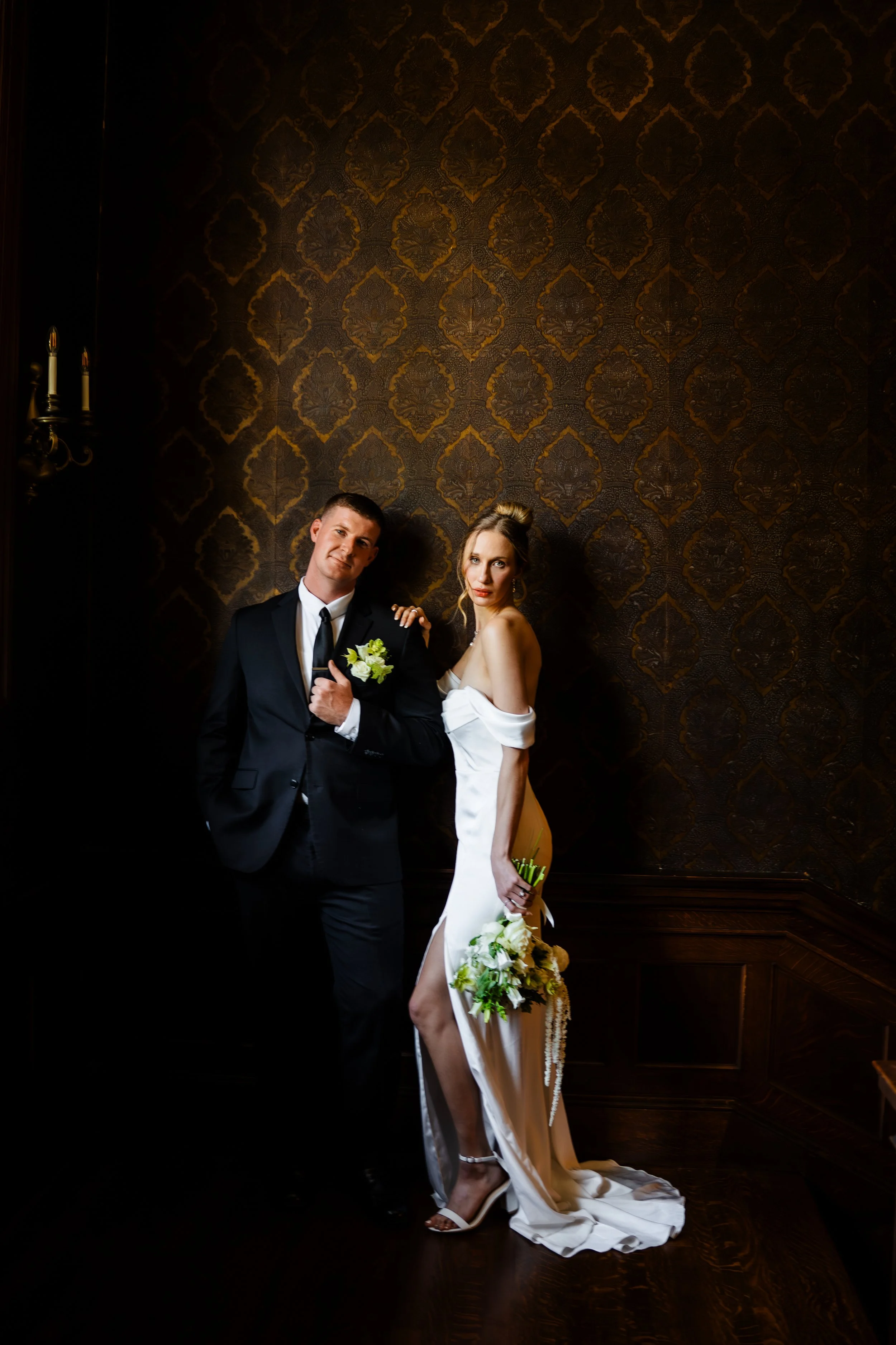 Bride and groom posing together in formal wedding attire against a dramatic dark editorial backdrop.
